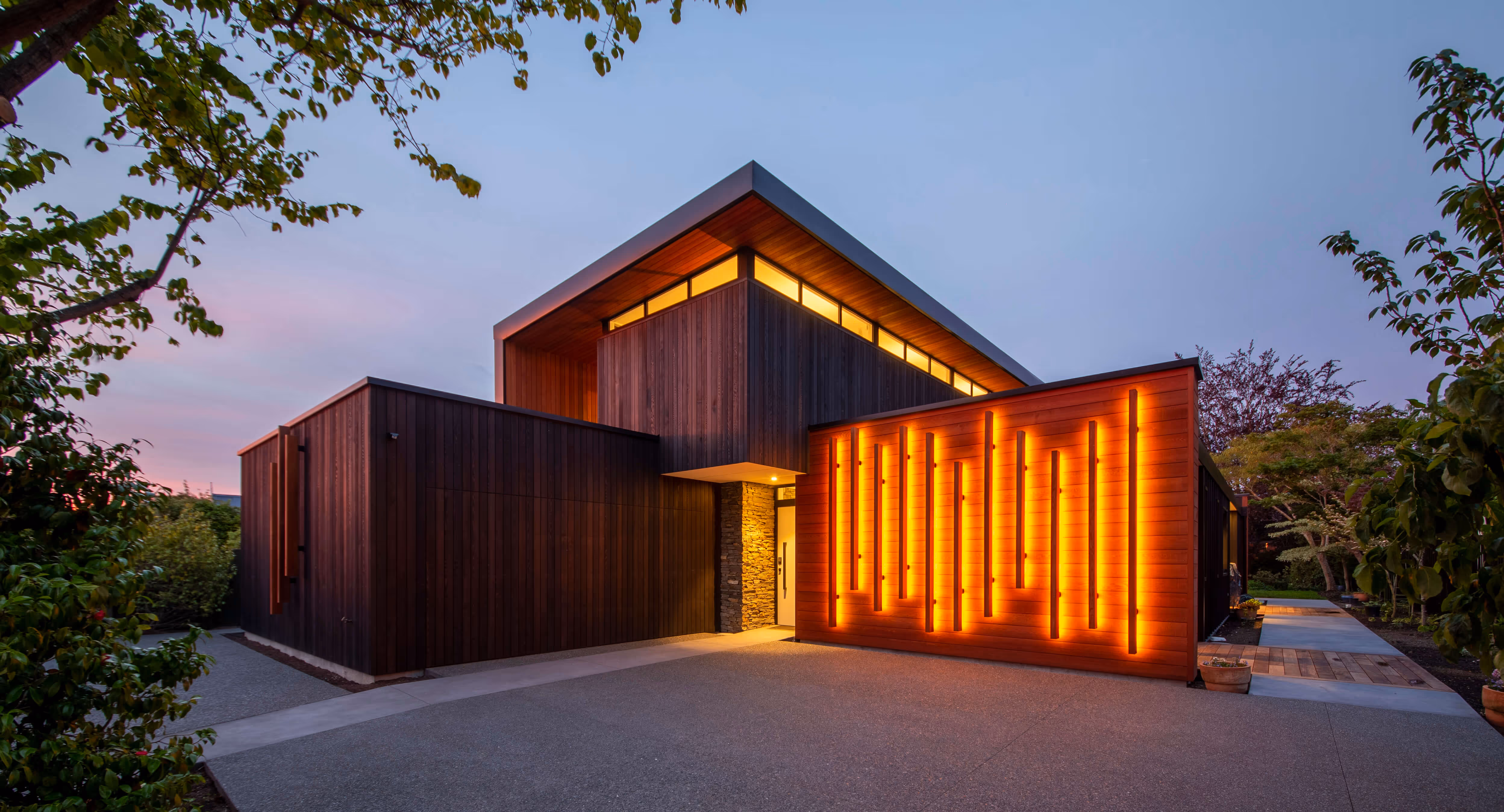 Modern dark wood house exterior at dusk with vertical warm orange light strips on one wall.