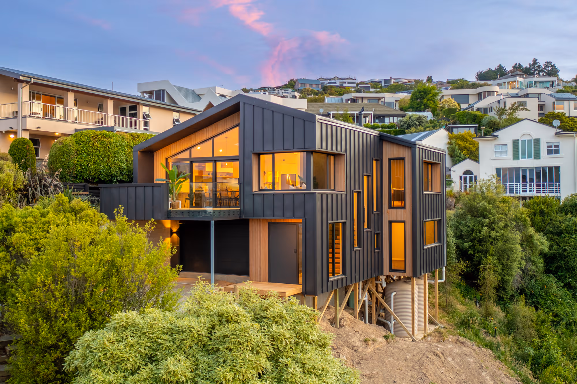 Modern black house with large windows and warm interior lights, built on stilts on a hillside with green bushes.