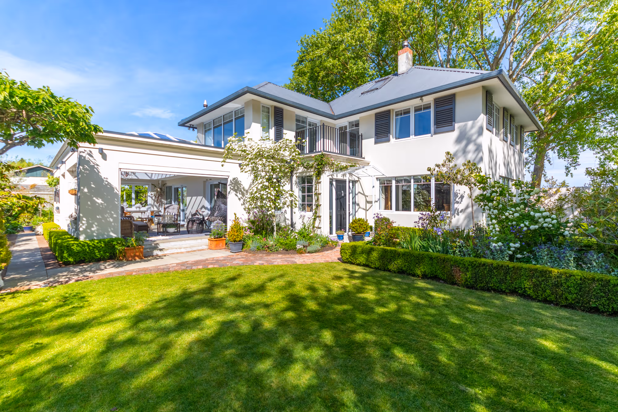Two-story white house with a gray roof, surrounded by lush green trees, bushes, and a well-maintained lawn under a blue sky.
