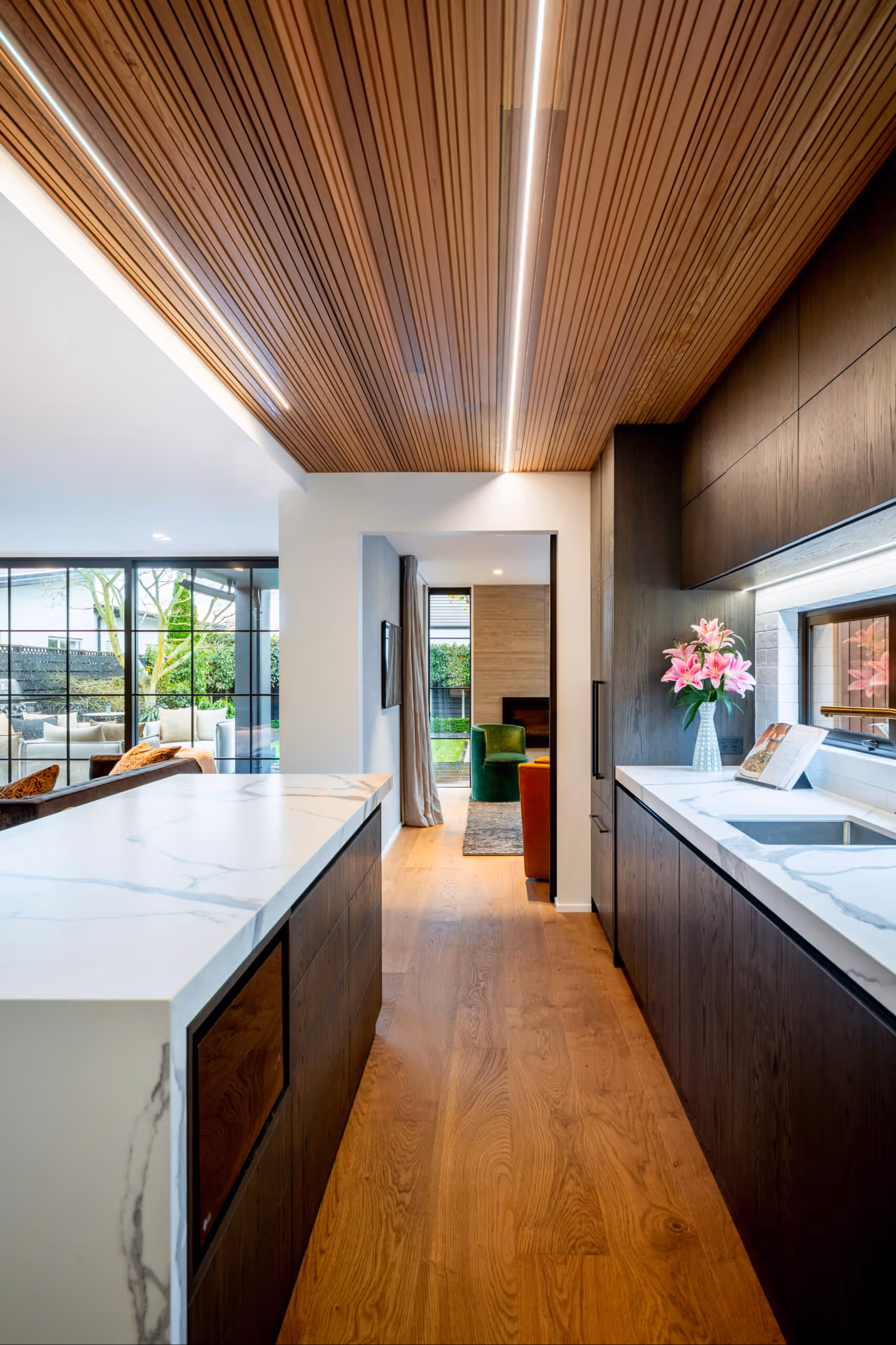 Modern kitchen with wooden floor and ceiling, white marble countertops, dark wood cabinets, a vase of pink flowers, and a view into a living room with large windows and green chairs.