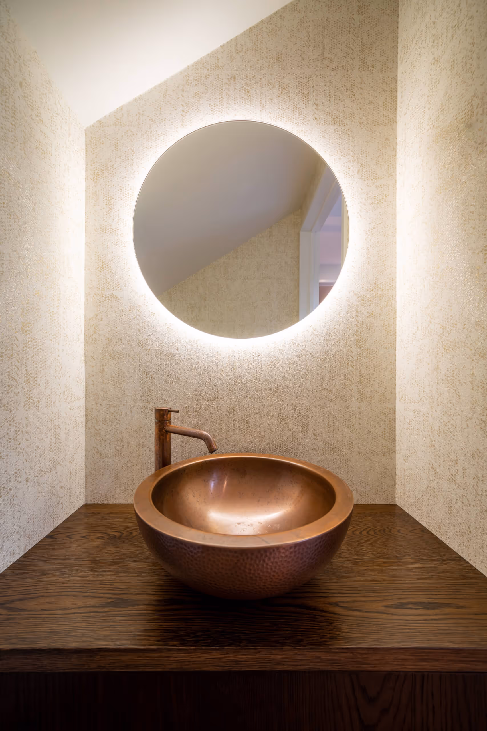 Modern bathroom vanity with a round copper sink, bronze faucet, and illuminated circular mirror on textured beige wall.