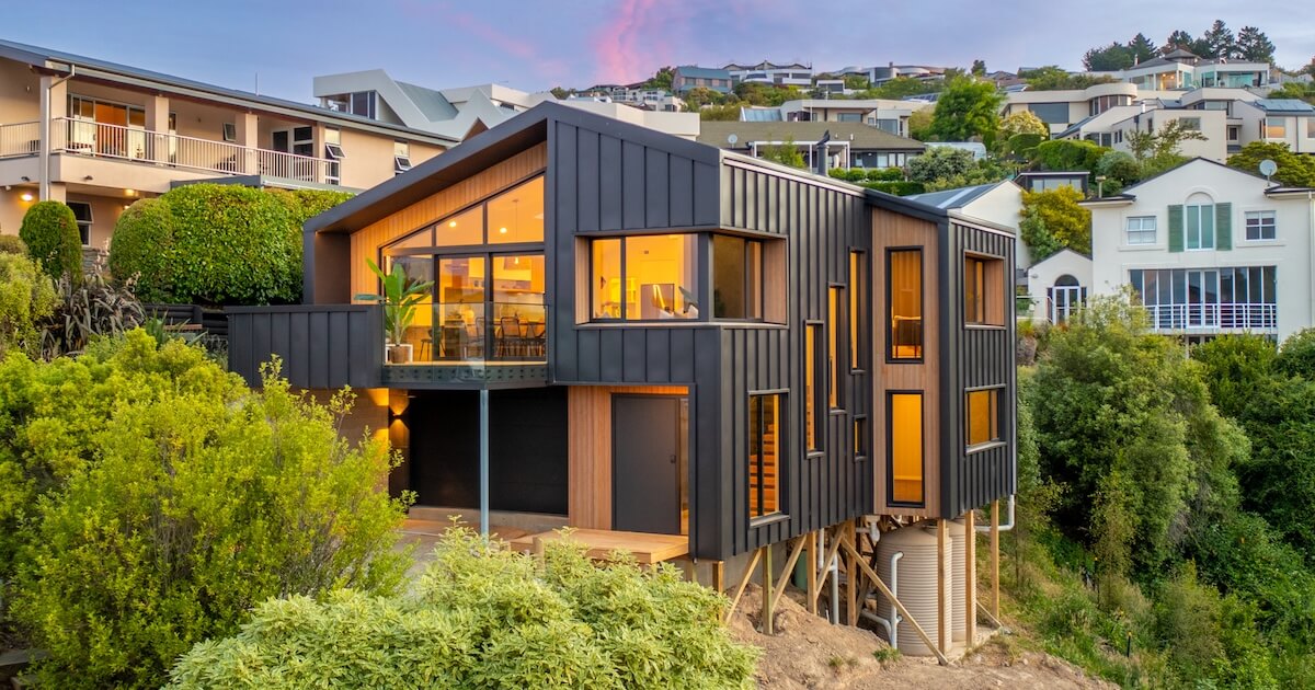 Modern house with large windows and warm interior lighting, elevated on wooden stilts on a hillside surrounded by greenery.