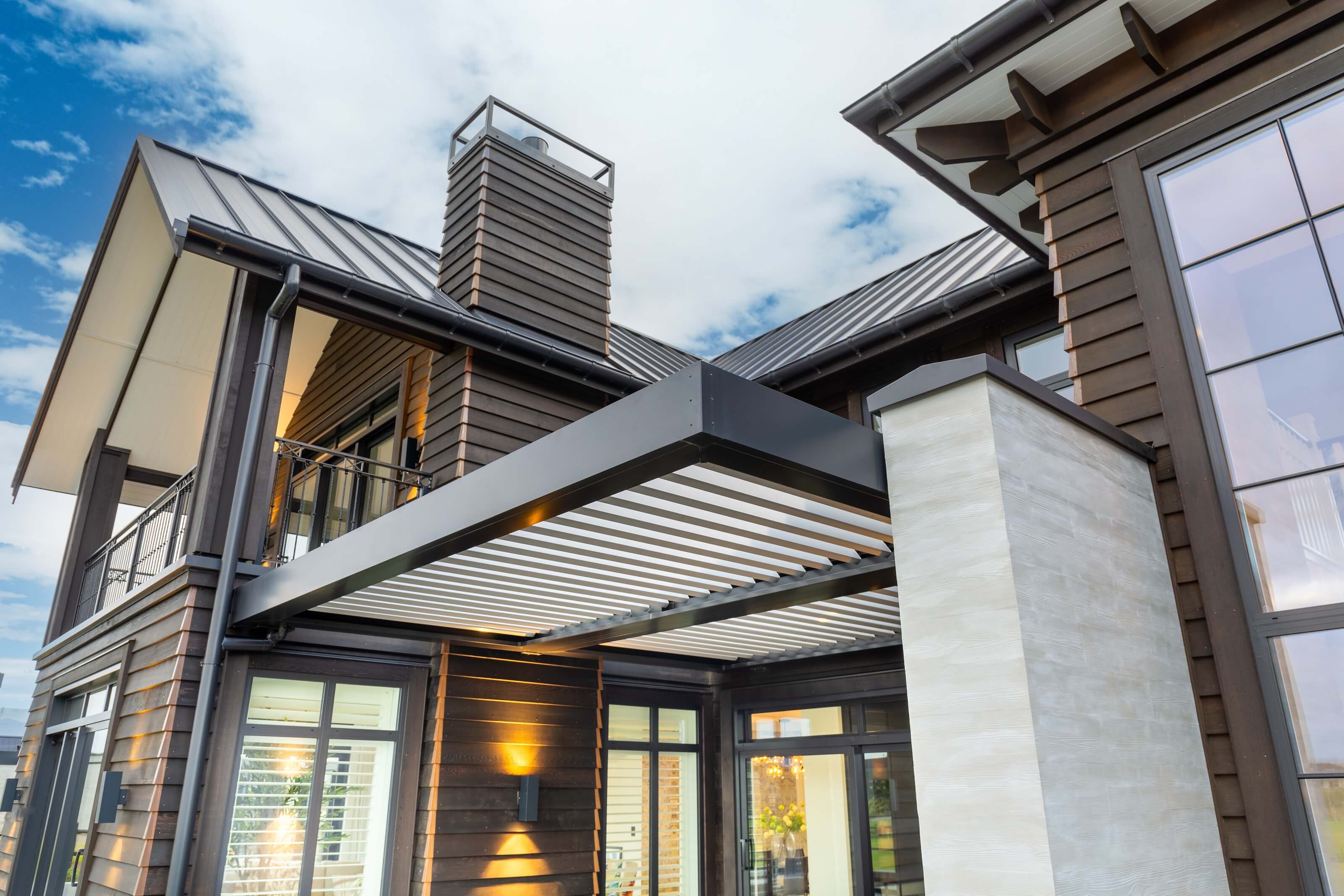 Modern house exterior with dark wood siding, large windows, a chimney, and a black metal pergola.