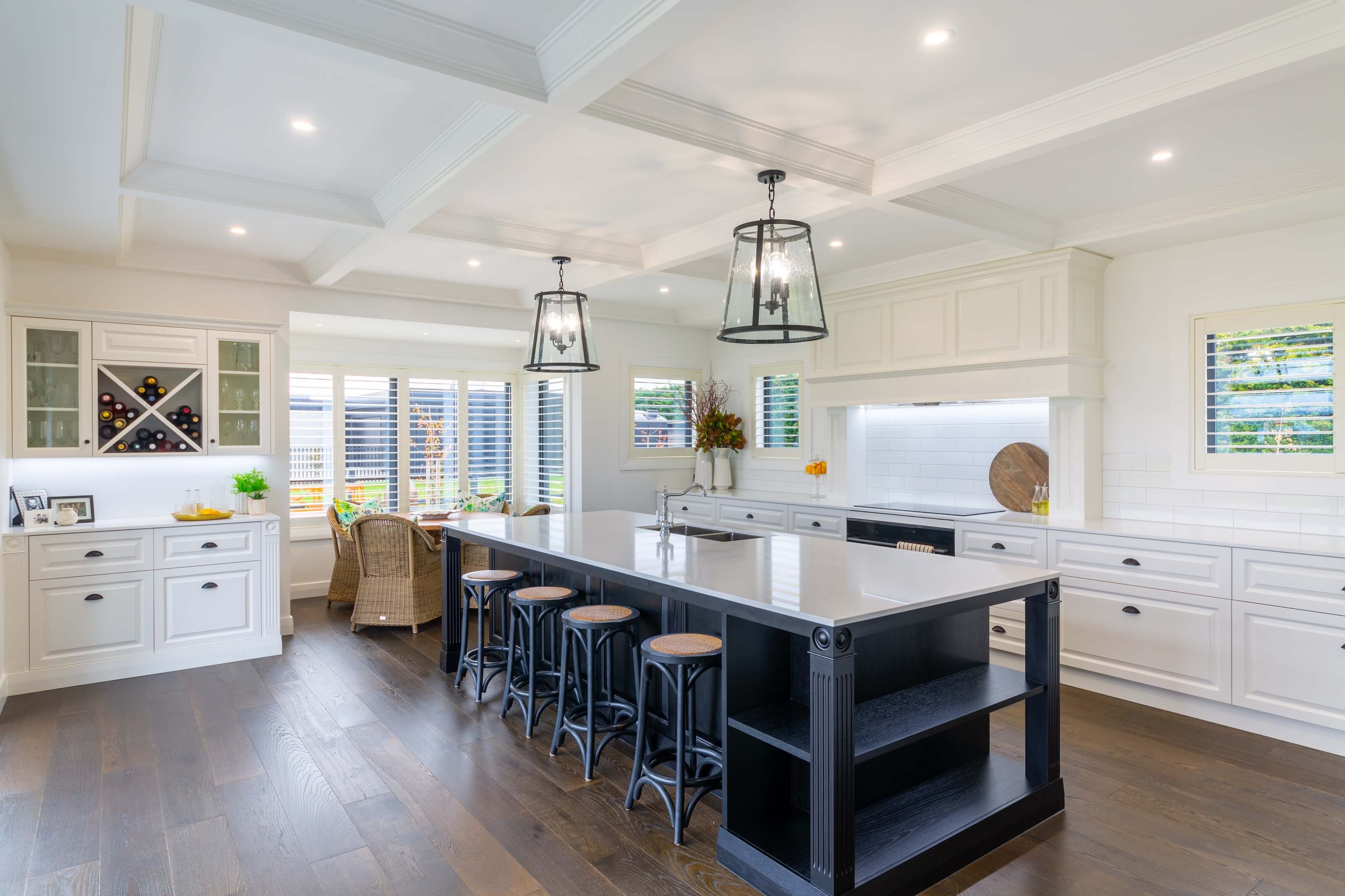 Bright kitchen with a large black island and white countertop, six black stools, white cabinetry, glass wine rack, wicker chairs by windows, and two hanging pendant lights.