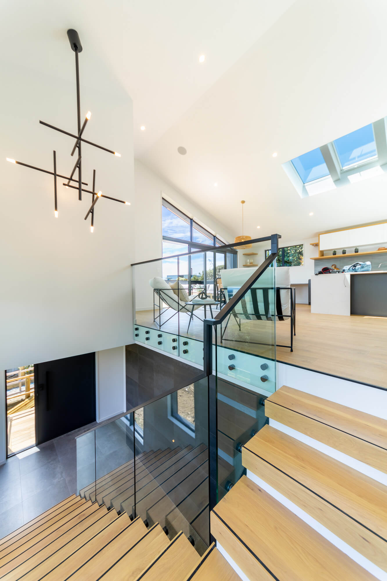 Modern staircase with glass railing and wooden steps leading to a bright living area with large windows and minimalist furniture.