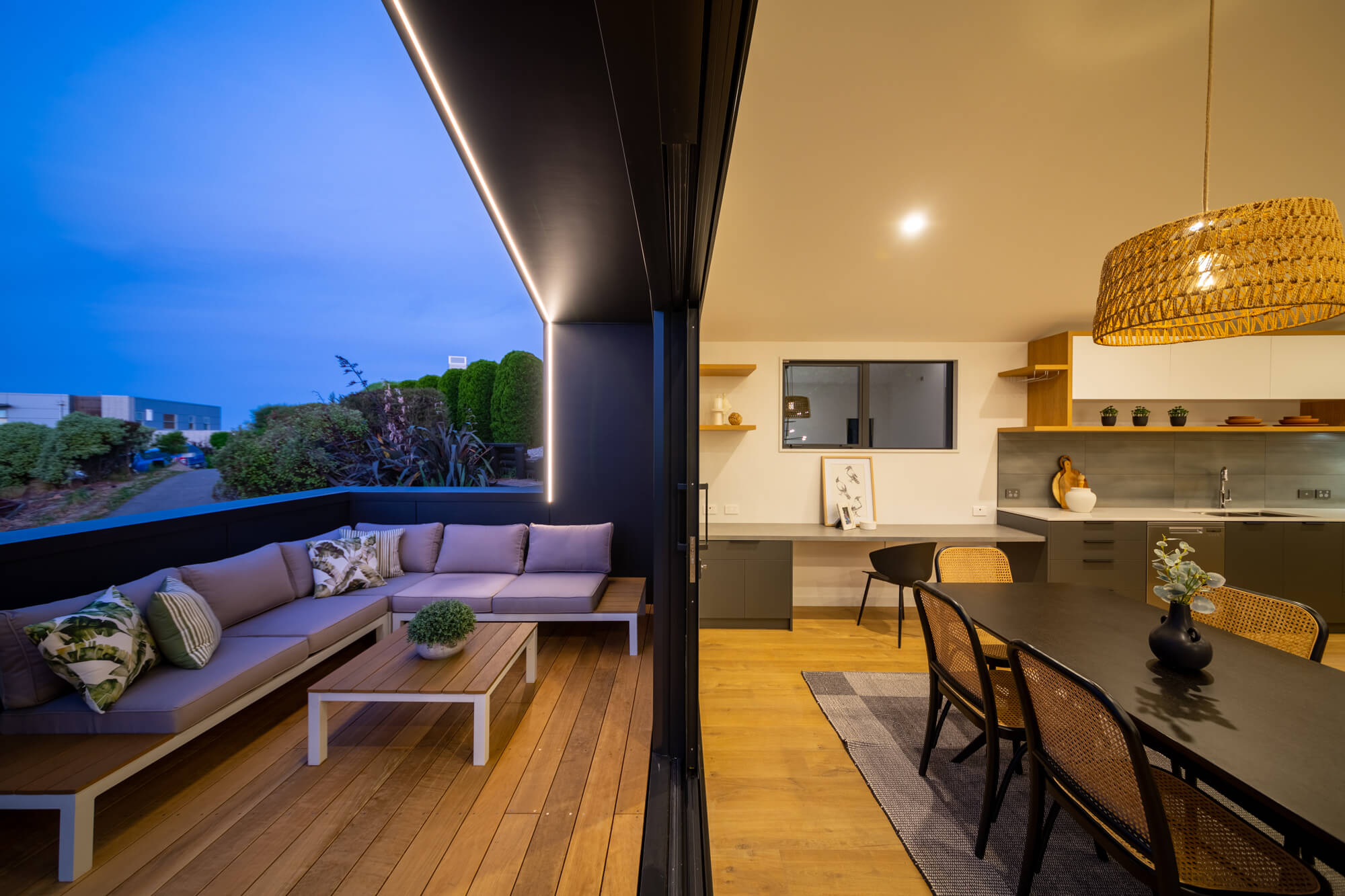 Modern outdoor patio with an L-shaped sofa and wooden coffee table, adjacent to an indoor dining area with black table, wicker chairs, and a hanging lamp.