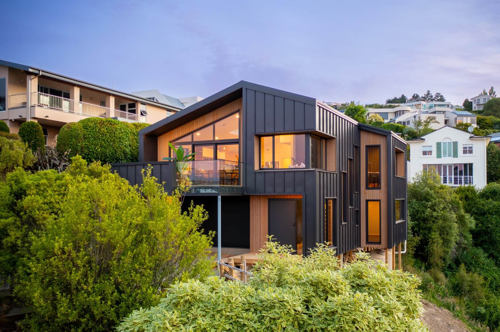 Modern two-story house with black paneling and large windows surrounded by green bushes at dusk.