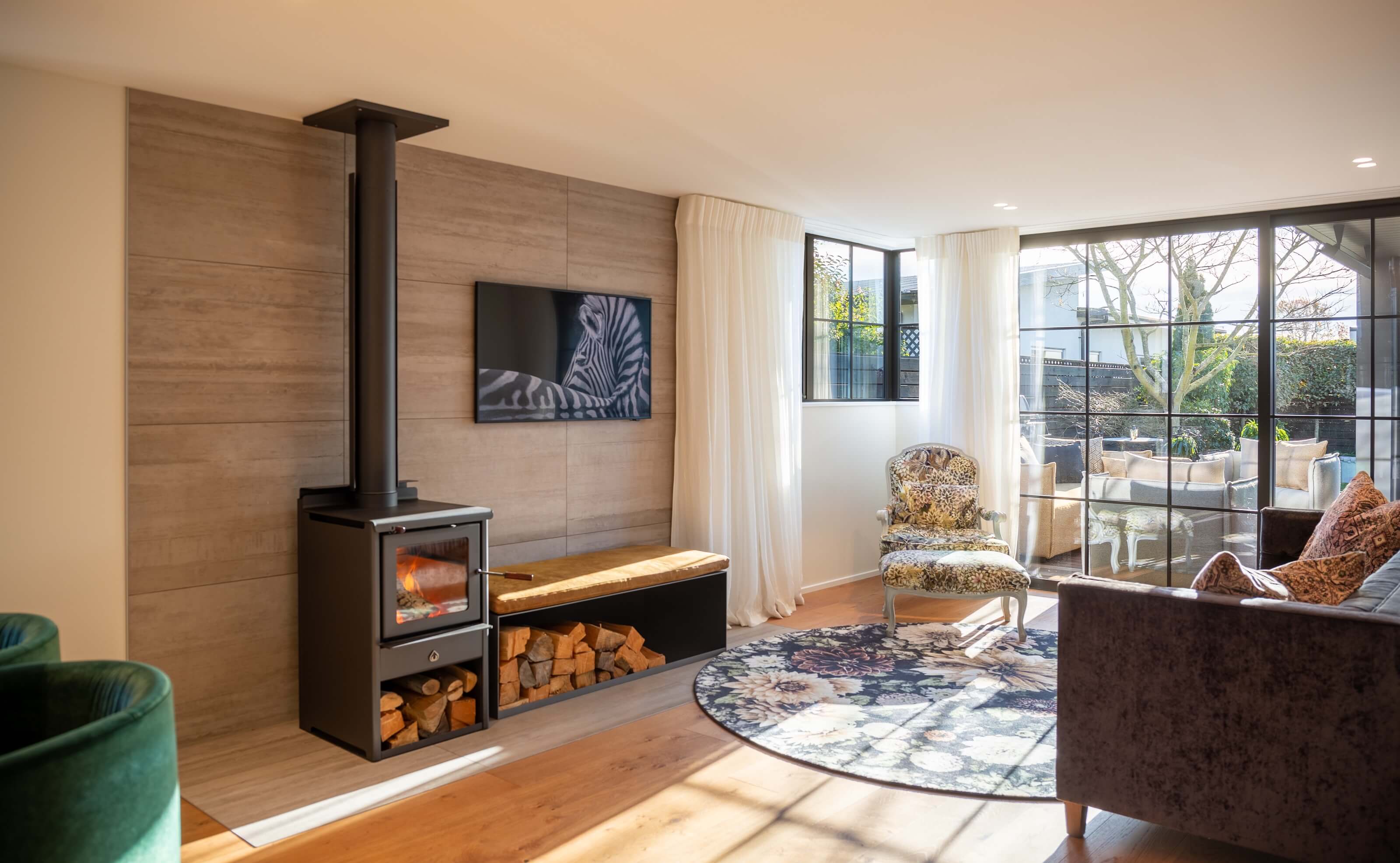 Modern living room with wood-burning stove, floral rug, vintage armchair, velvet sofa, and large glass doors opening to an outdoor seating area.