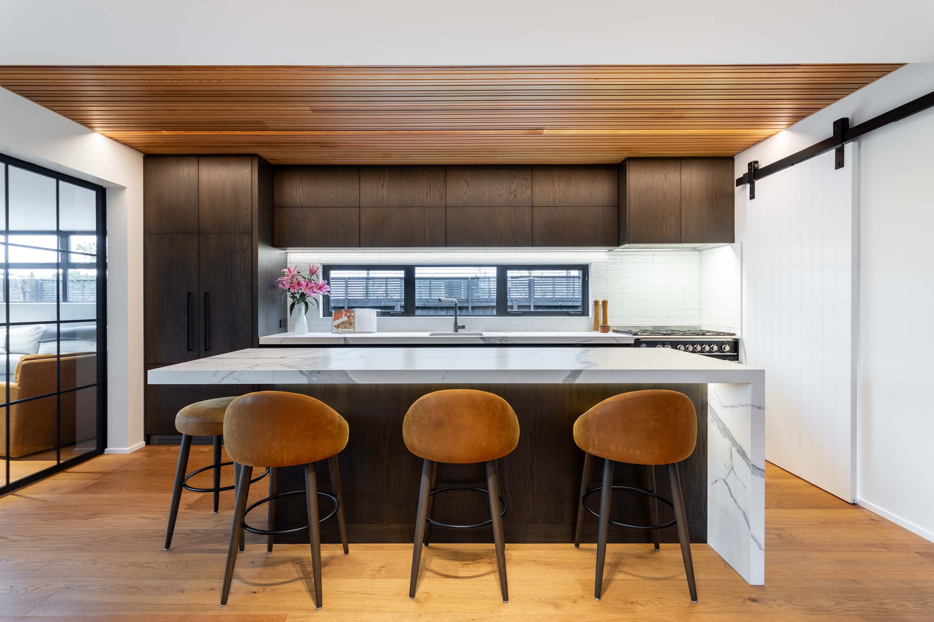Modern kitchen with white marble island, four brown velvet bar stools, dark wood cabinets, and a wooden slatted ceiling.
