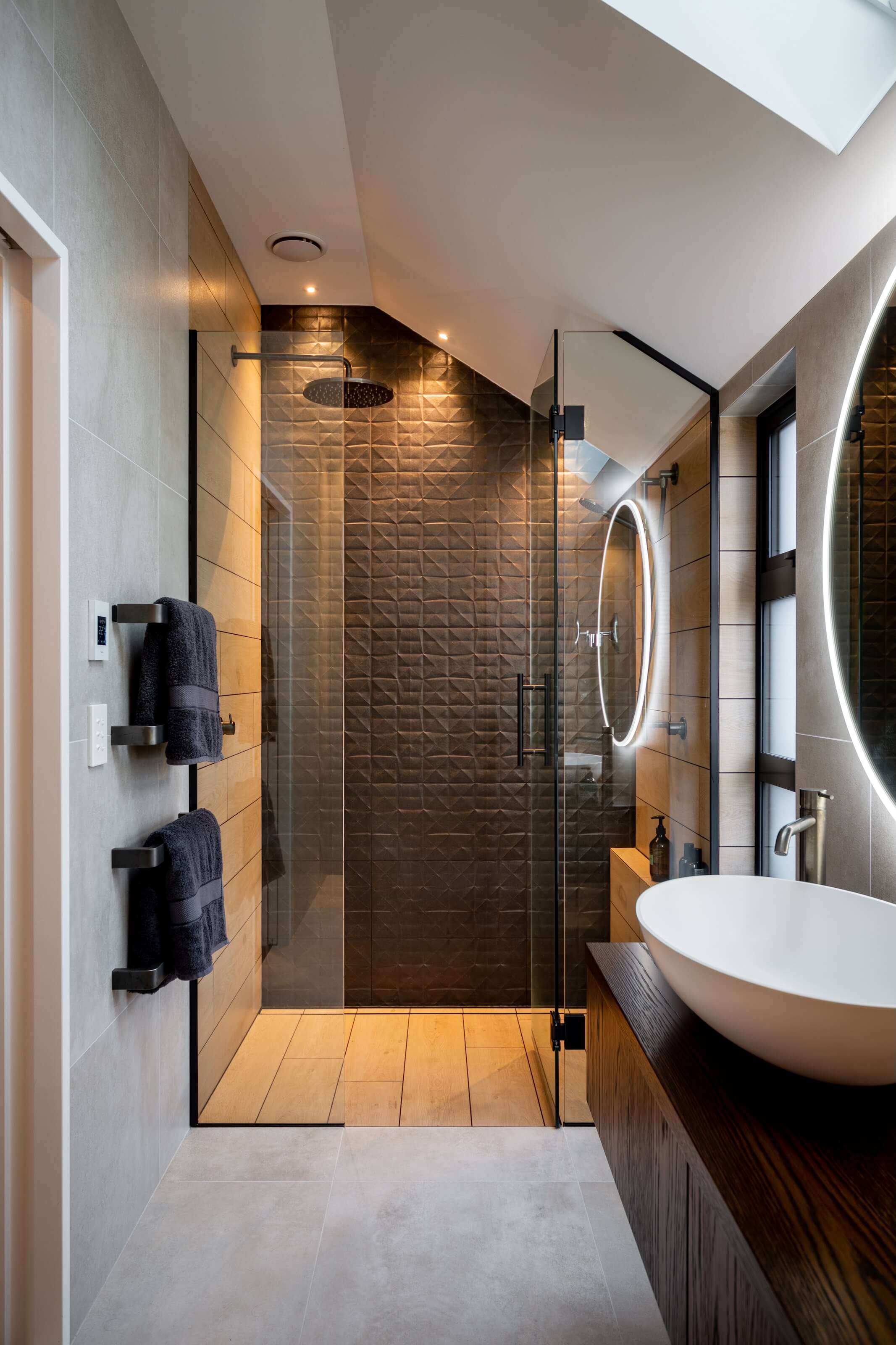 Modern bathroom with a glass-enclosed shower featuring dark textured tiles, wood-look floor tiles, and a vanity with a round illuminated mirror and a white vessel sink.