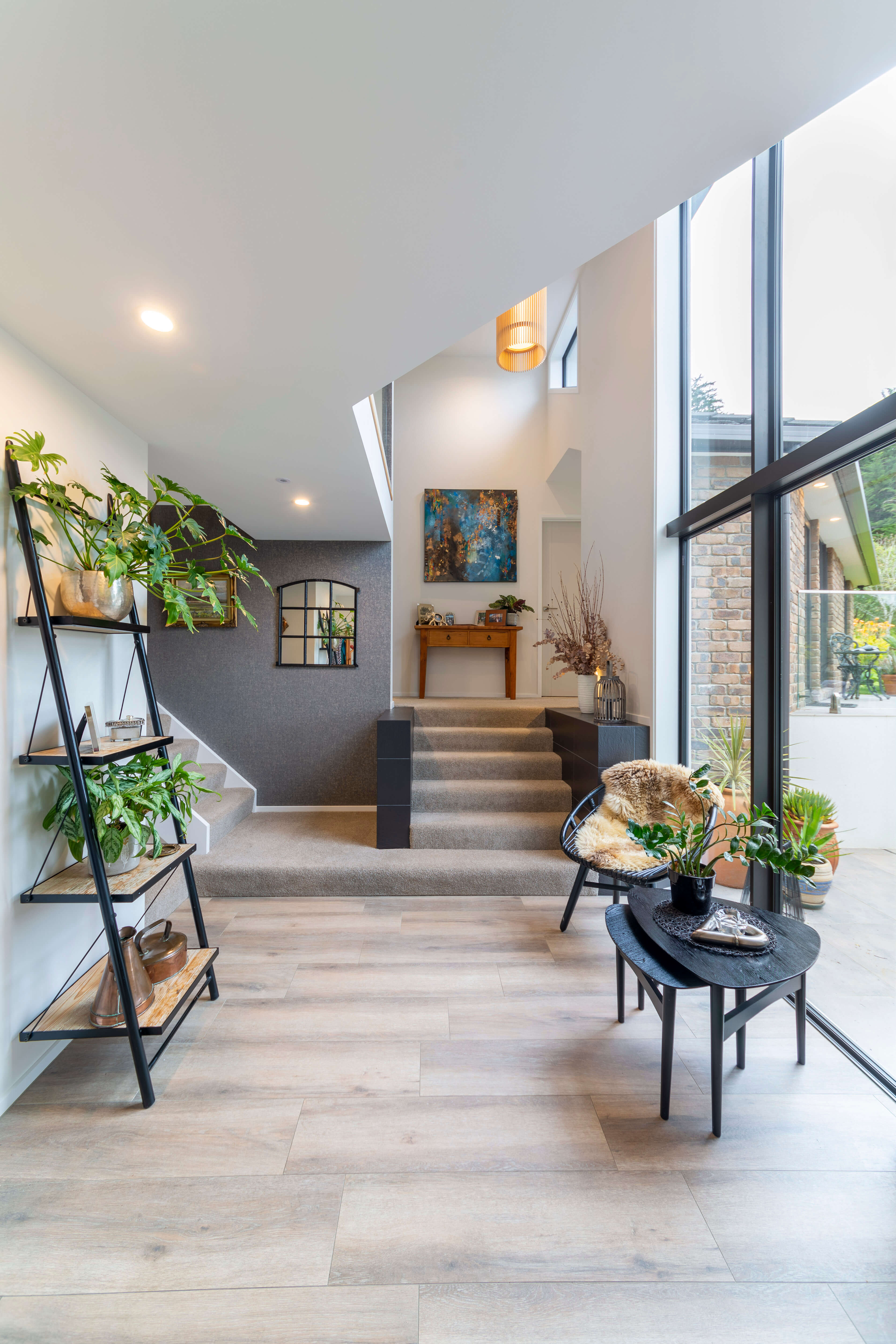 Bright modern entryway with wooden flooring, carpeted stairs, black metal shelves with plants, a black chair with fur throw, and large windows overlooking a patio.