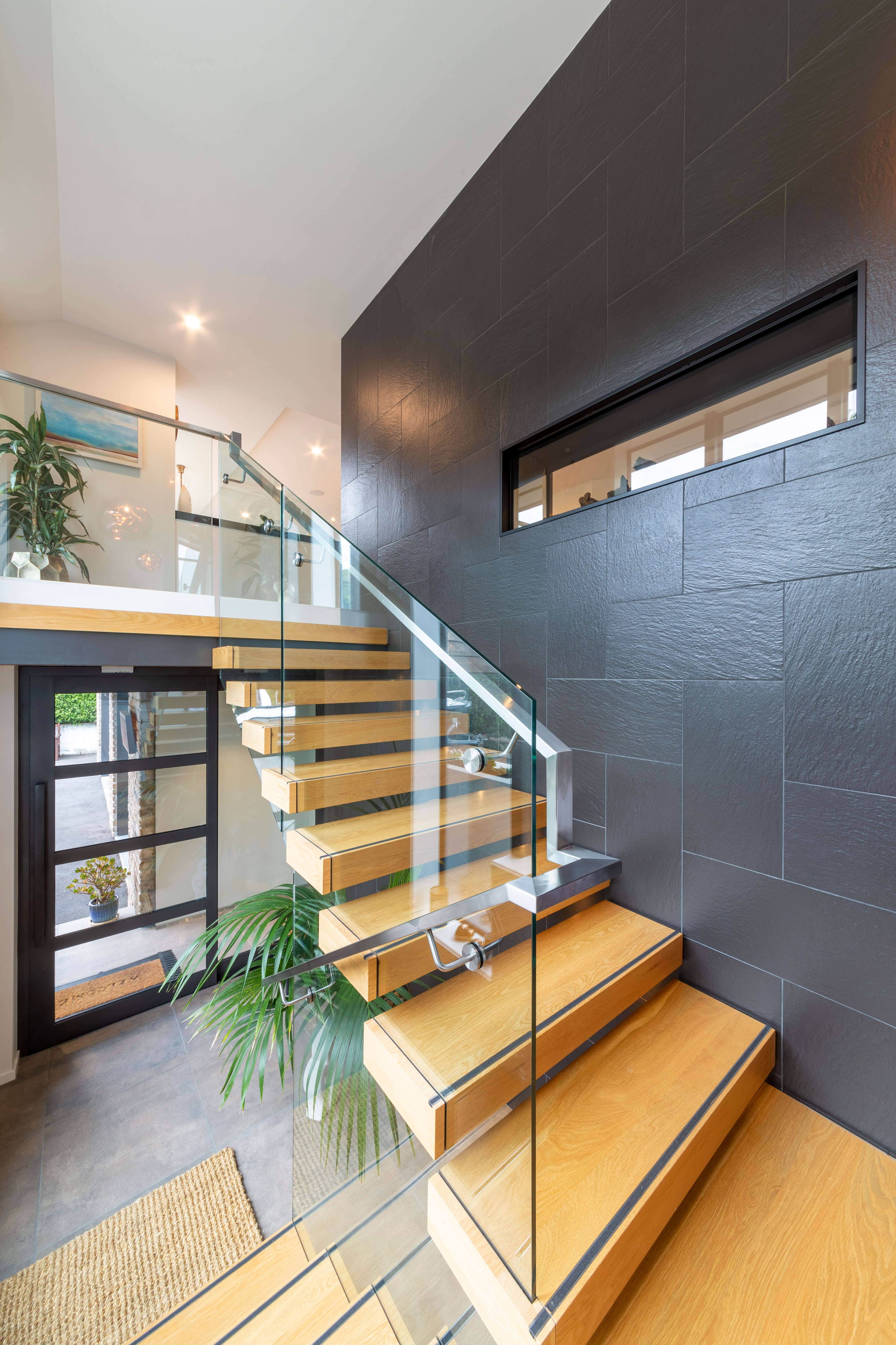 Modern interior with wooden floating staircase featuring glass railing and a dark tiled accent wall.