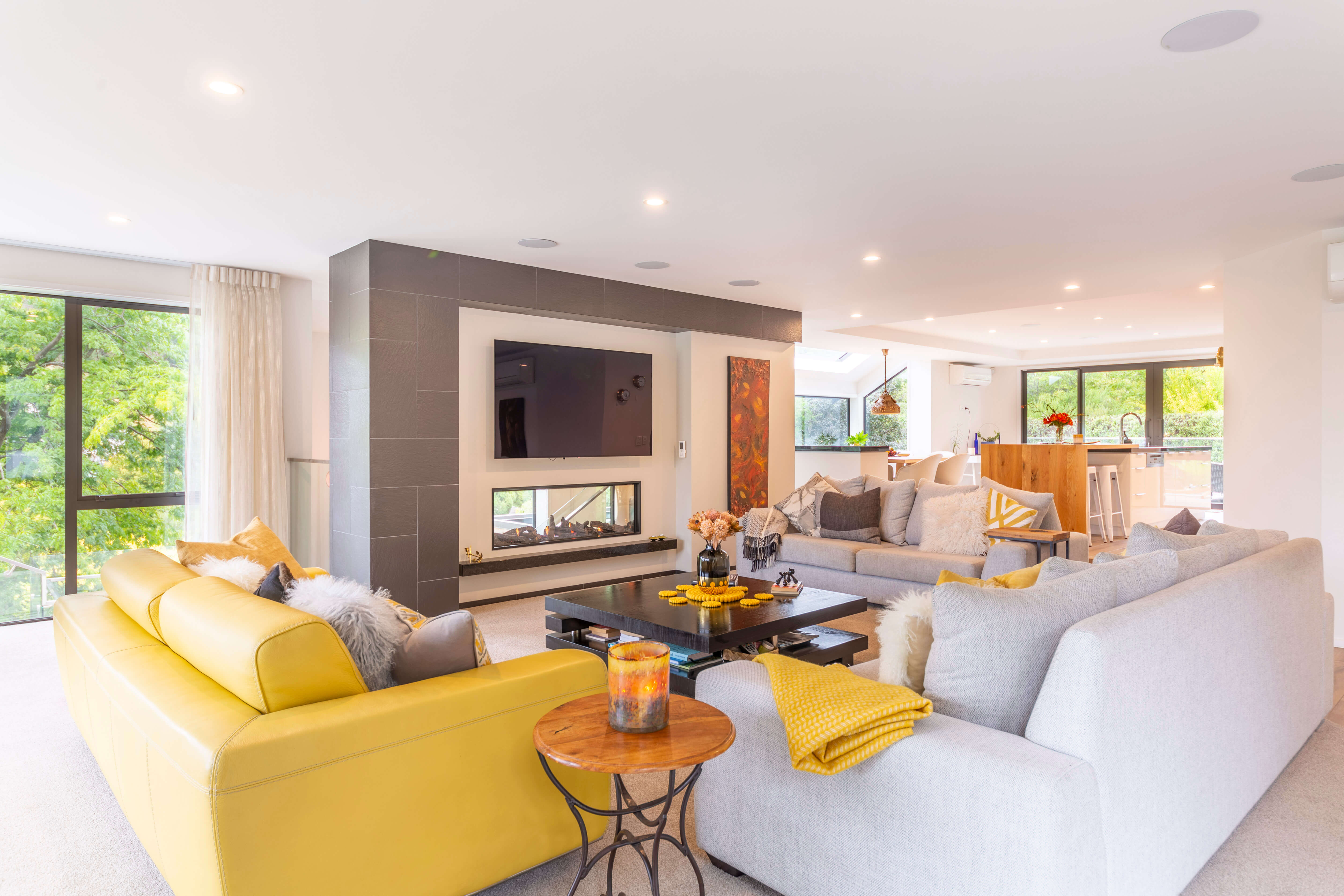 Bright modern living room with yellow and gray sofas, a black coffee table, fireplace, and large windows showing green trees outside.