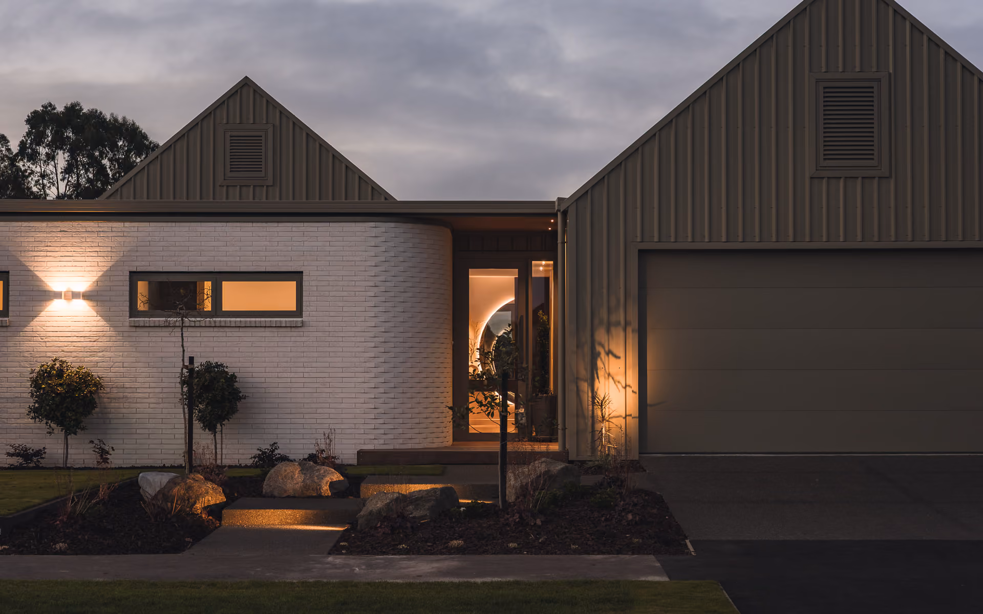 Modern house exterior at dusk with lit entrance, white brick wall, and closed garage door.