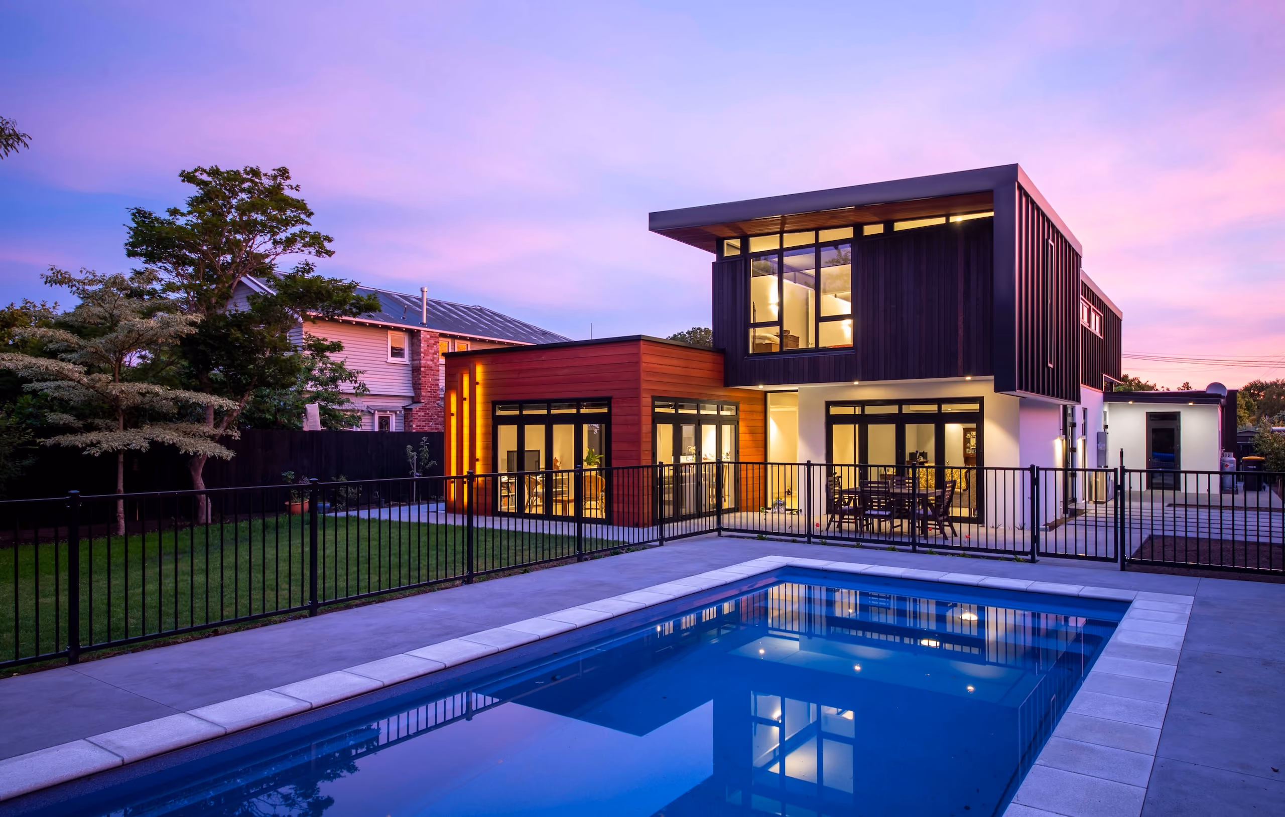 Modern two-story house with wooden accents, large windows, patio, and a fenced pool at dusk.