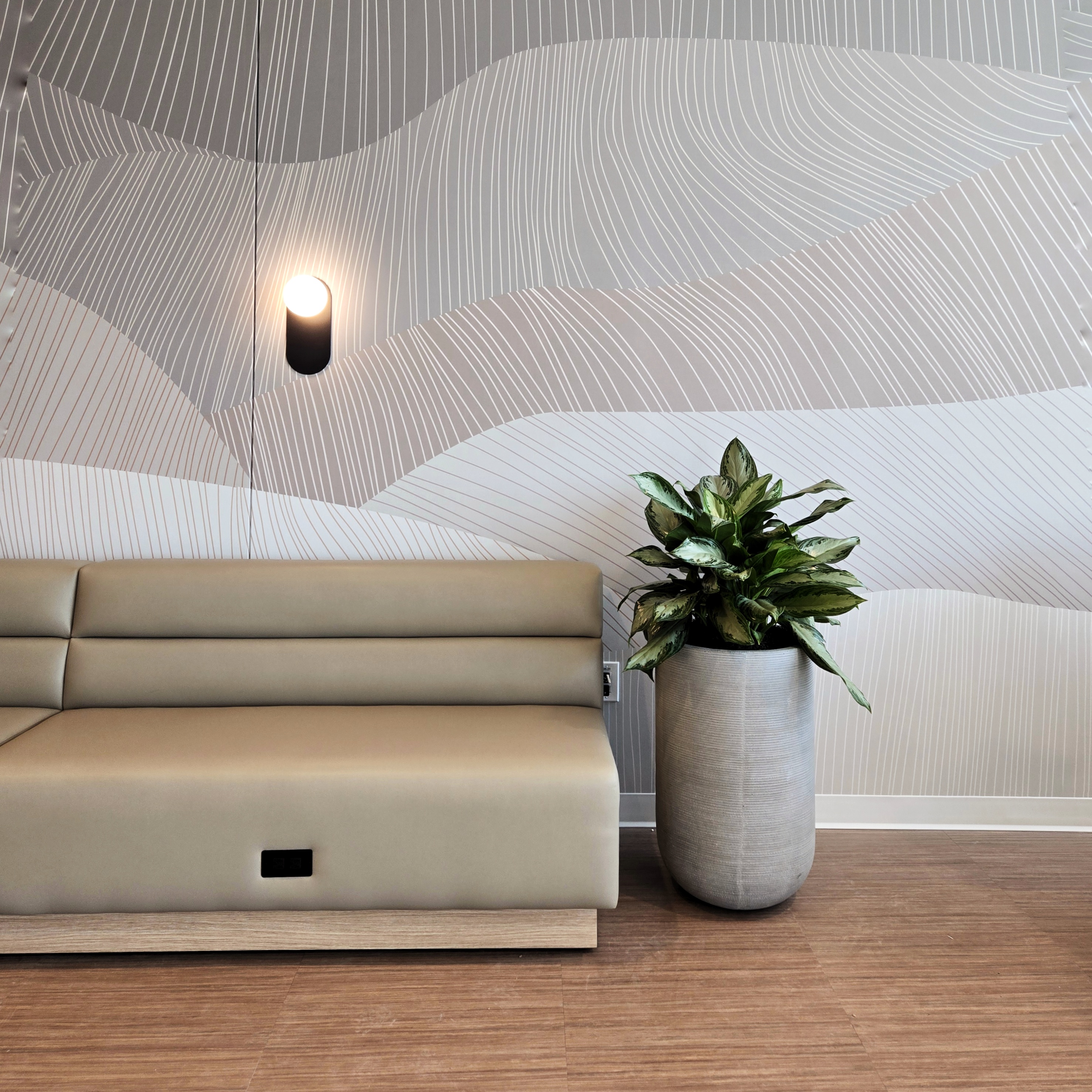 Modern beige bench with a power outlet, beside a large cylindrical planter holding a green leafy plant, set against a wall with abstract curved line patterns in neutral tones and a single wall-mounted cylindrical light fixture.