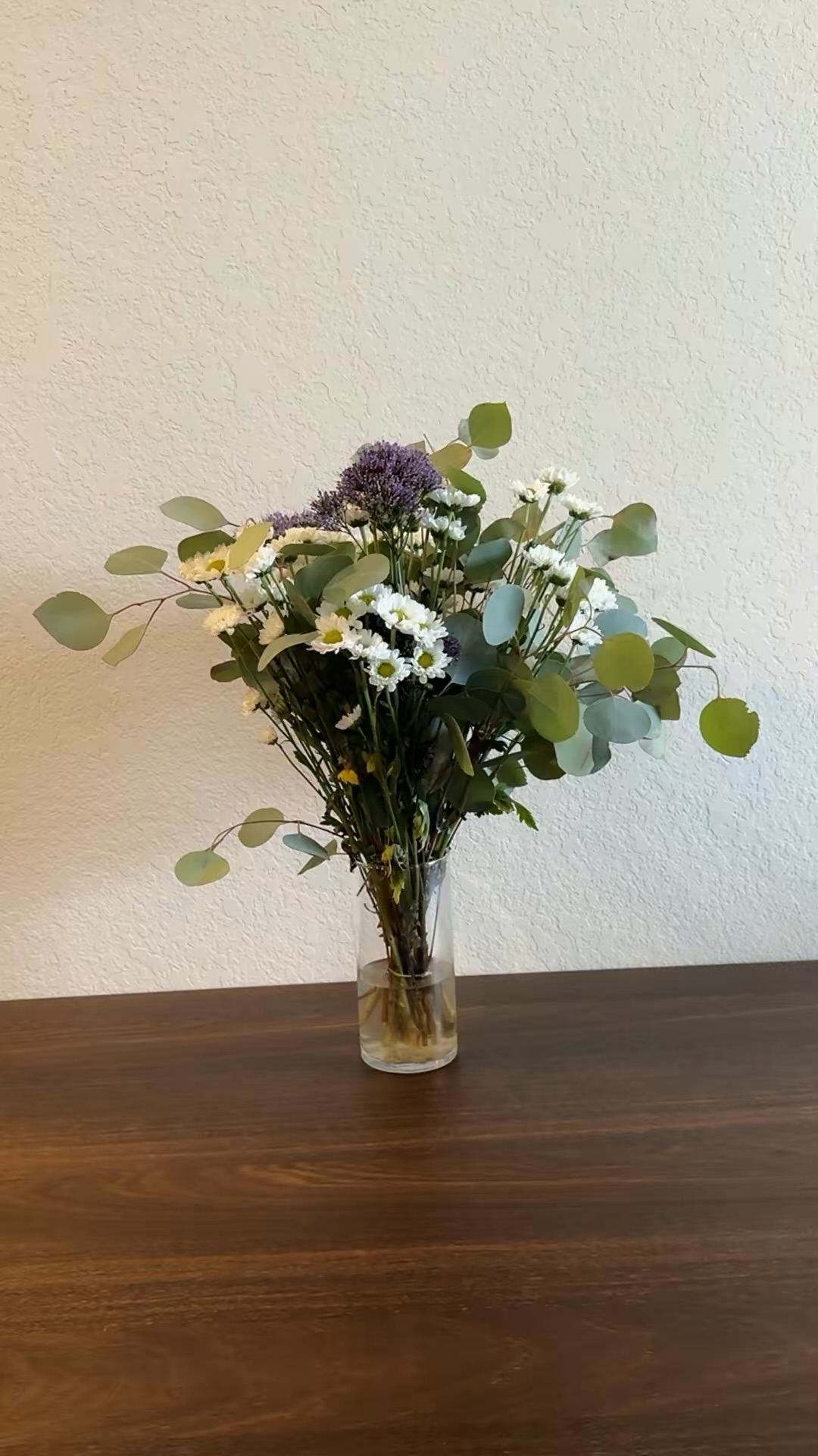Clear glass vase with a bouquet of white daisies, green eucalyptus leaves, and purple flowers on a wooden table against a textured cream wall.