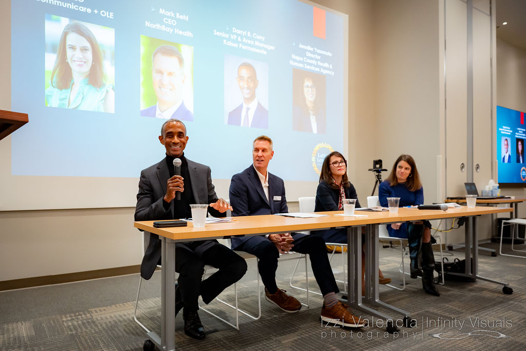 Four professionals seated at a panel table during a conference, with a large screen displaying speaker portraits and names behind them.