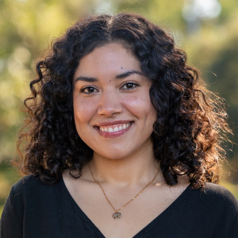 Smiling woman with curly dark hair and dimples, wearing a black top and a gold necklace with a round pendant, outdoors with blurred greenery background.