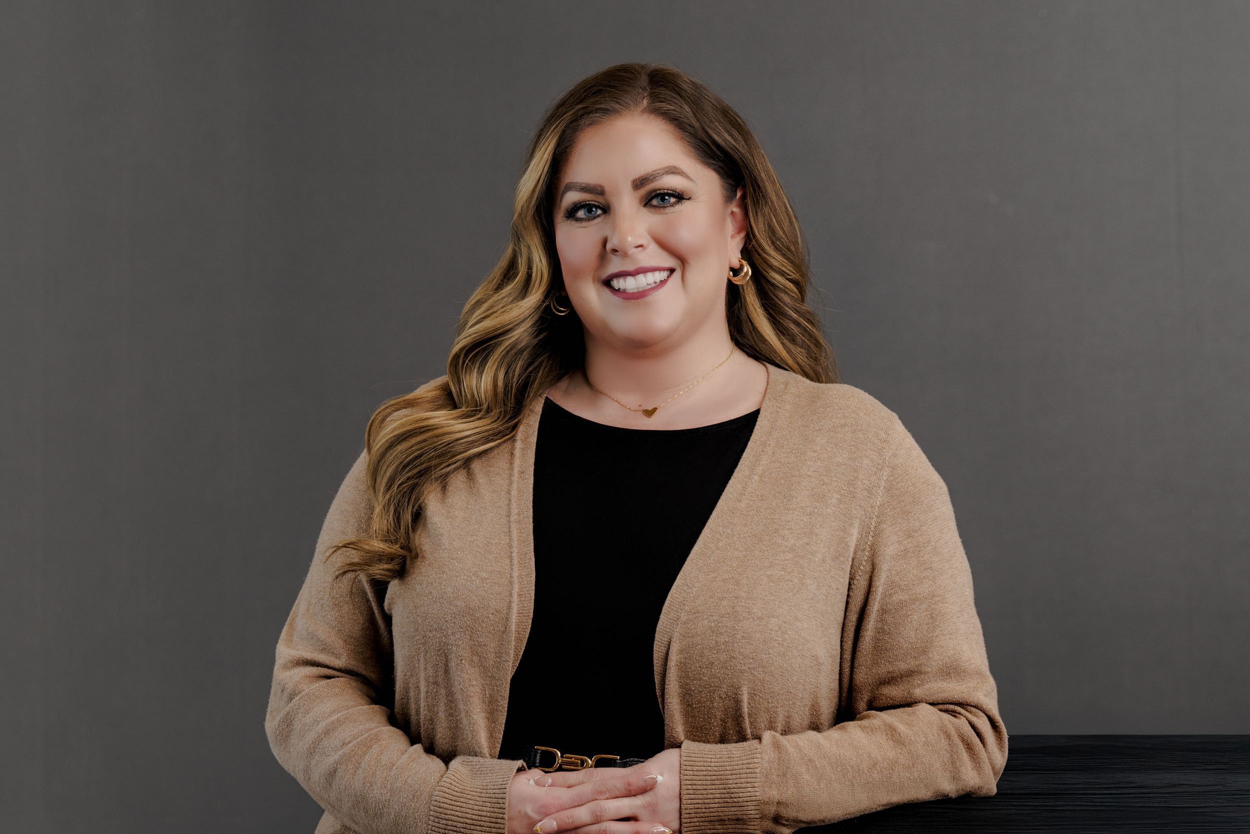 Woman with long wavy light brown hair wearing a black top and beige cardigan, smiling with hands clasped in front of a gray background.
