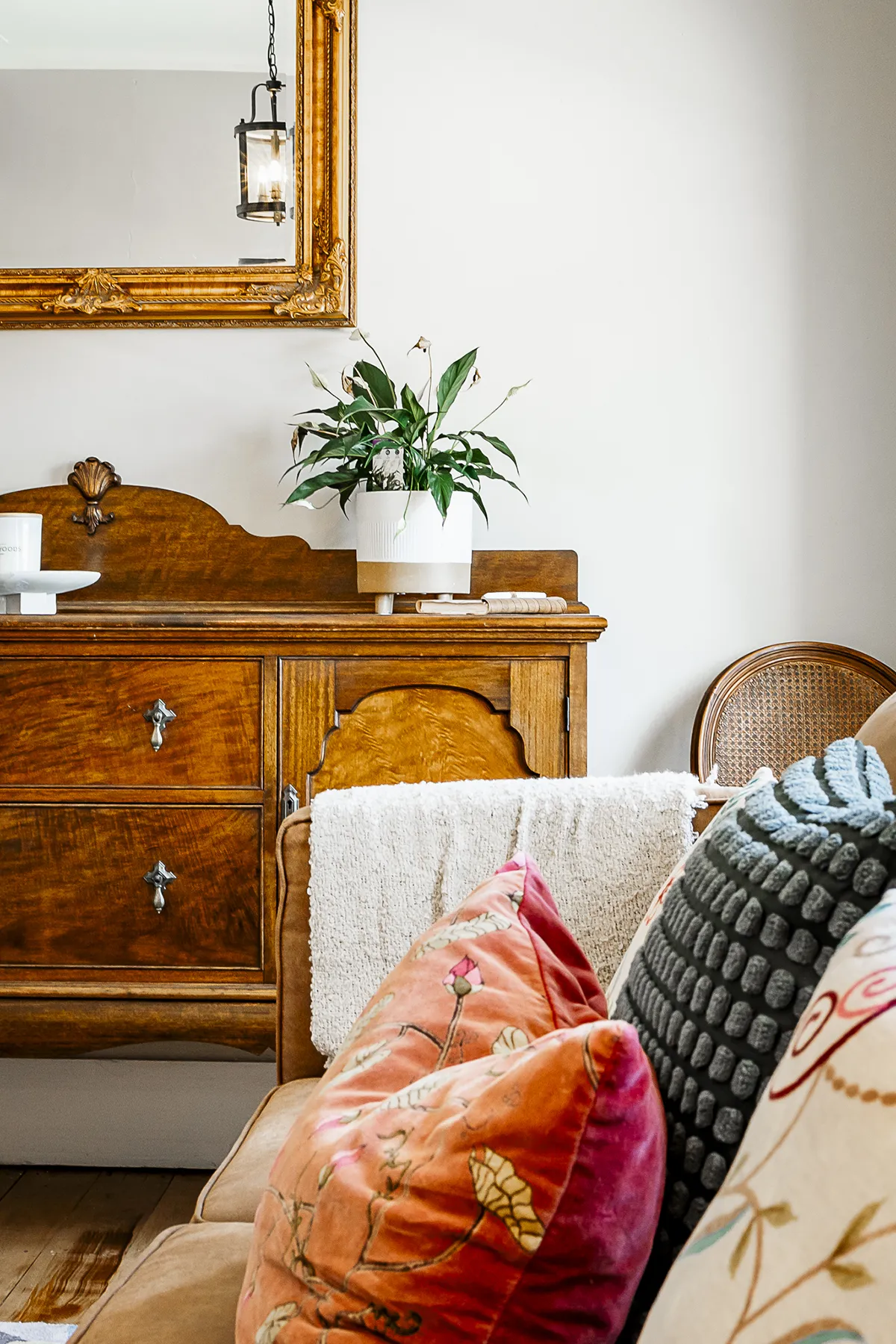 Cozy living room corner with wooden dresser holding a potted plant, large gold-framed mirror, and a beige sofa adorned with colorful patterned cushions and a white throw blanket.