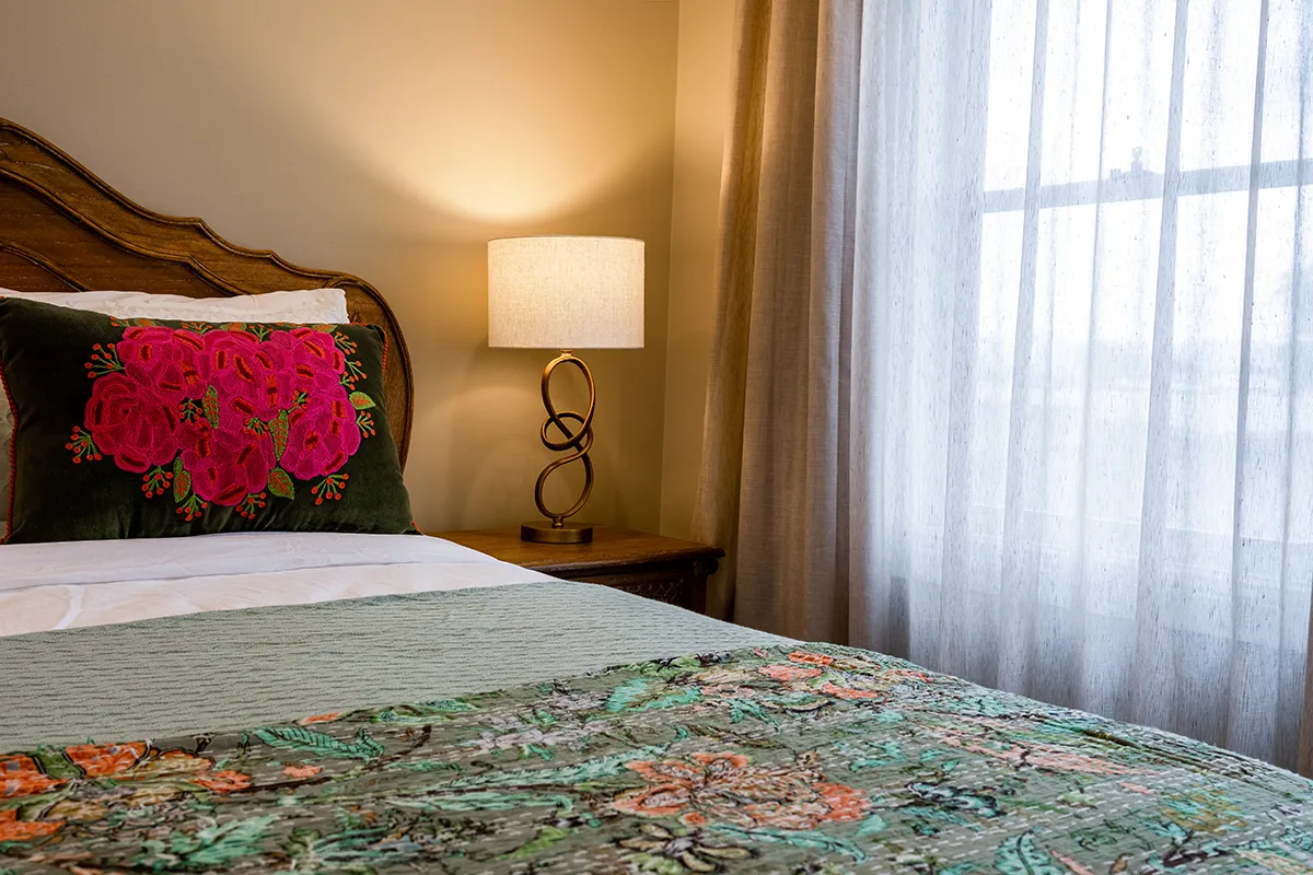Bedroom with a wooden headboard, a floral embroidered pillow, a green quilt, and a lit lamp on a wooden nightstand next to a window with sheer curtains.