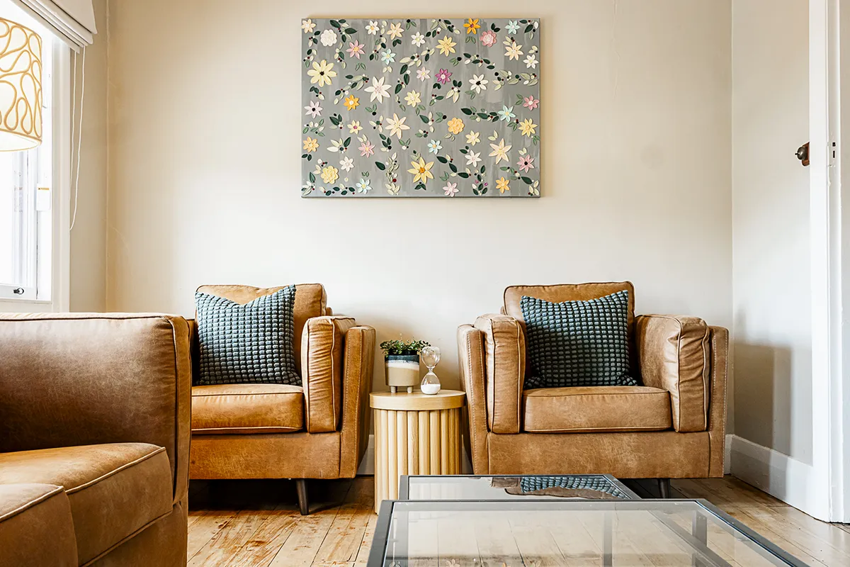 Cozy living room with two tan leather armchairs, blue textured pillows, a round wooden side table with a small plant and hourglass, and floral artwork on the wall.