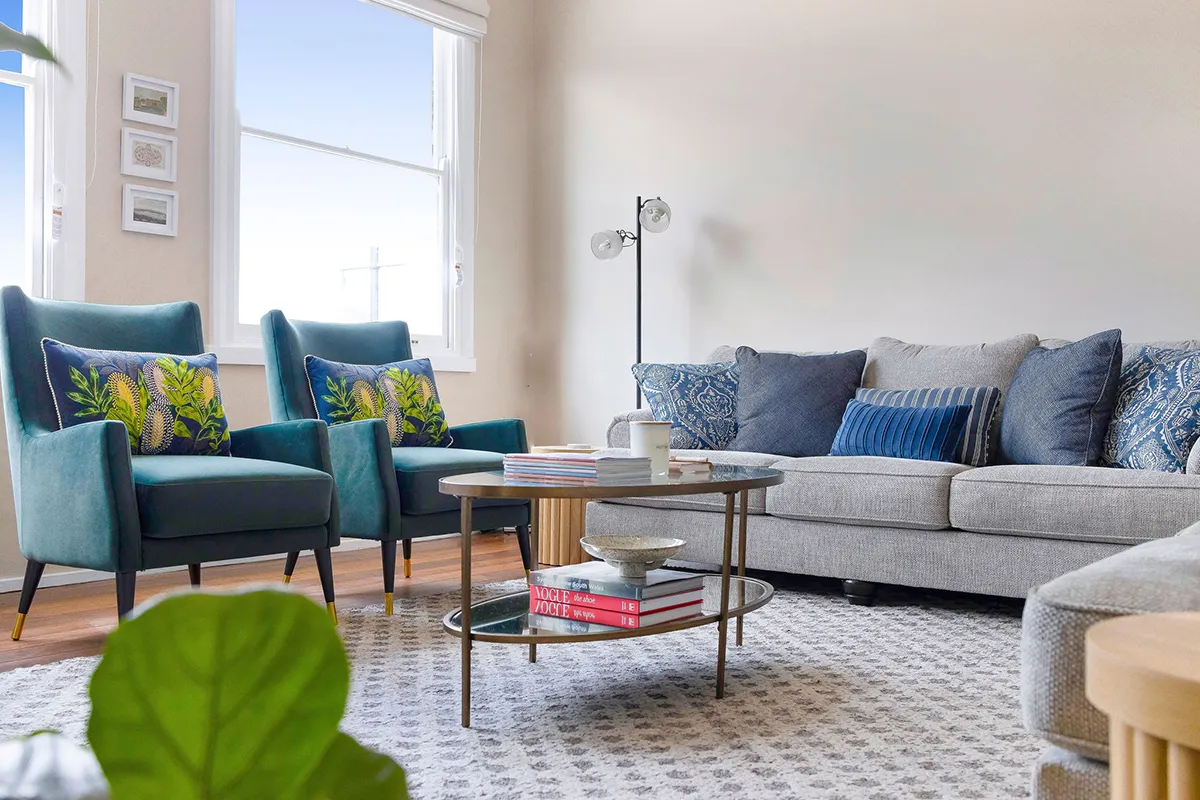 Modern living room with two teal armchairs with botanical cushions, a gray sofa with blue patterned pillows, and a round coffee table with books and a cup on a patterned rug.