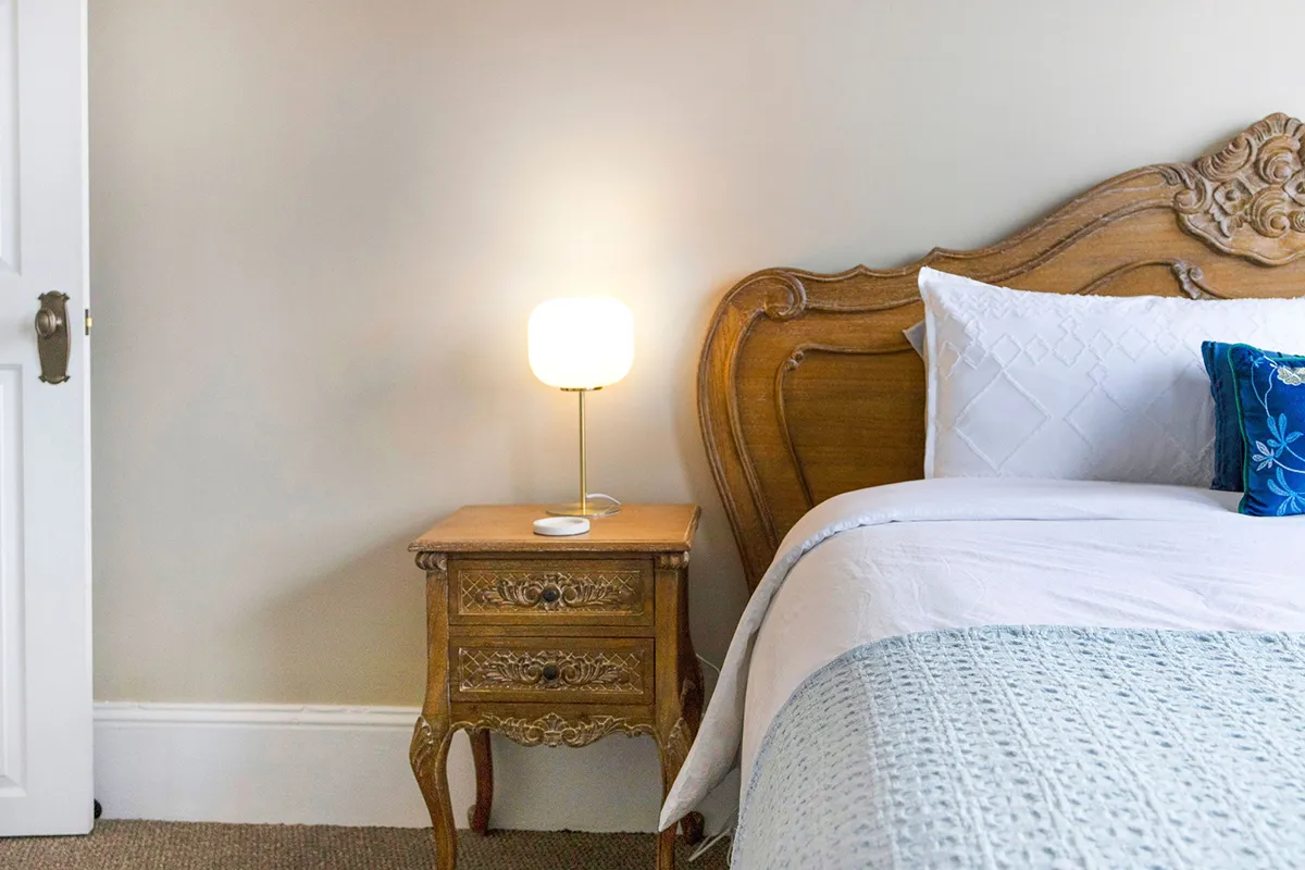 Wooden nightstand with ornate carvings beside a bed with a carved wooden headboard, white and blue pillows, and a lit table lamp on the nightstand.