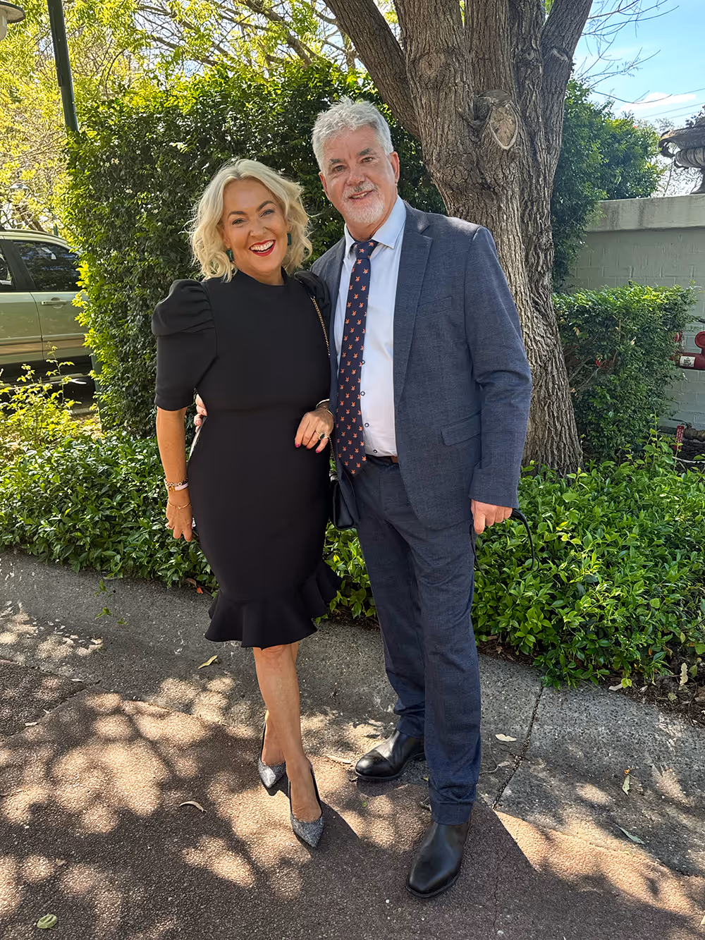 A smiling man in a blue suit and a woman in a black dress posing outdoors in front of a tree and greenery on a sunny day.