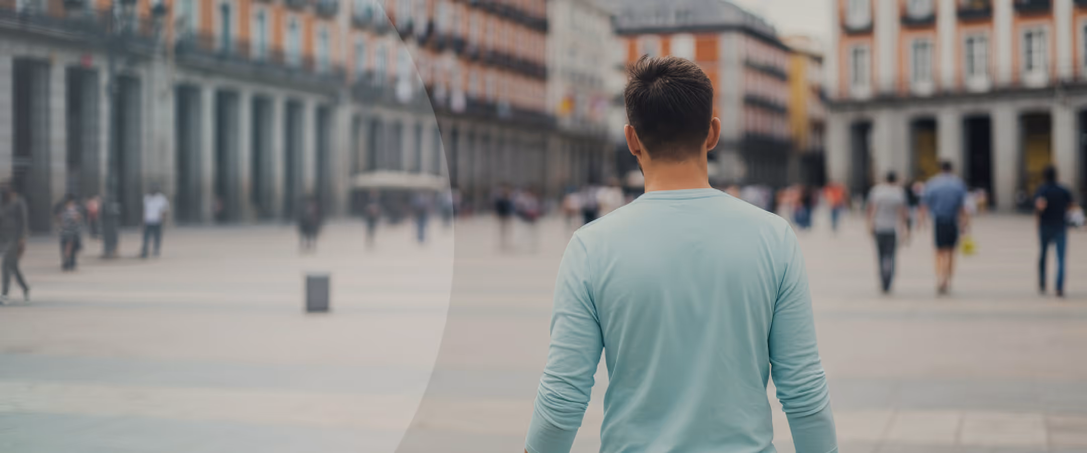 Man wearing a light blue long-sleeve shirt seen from behind, standing on a blurry urban street.