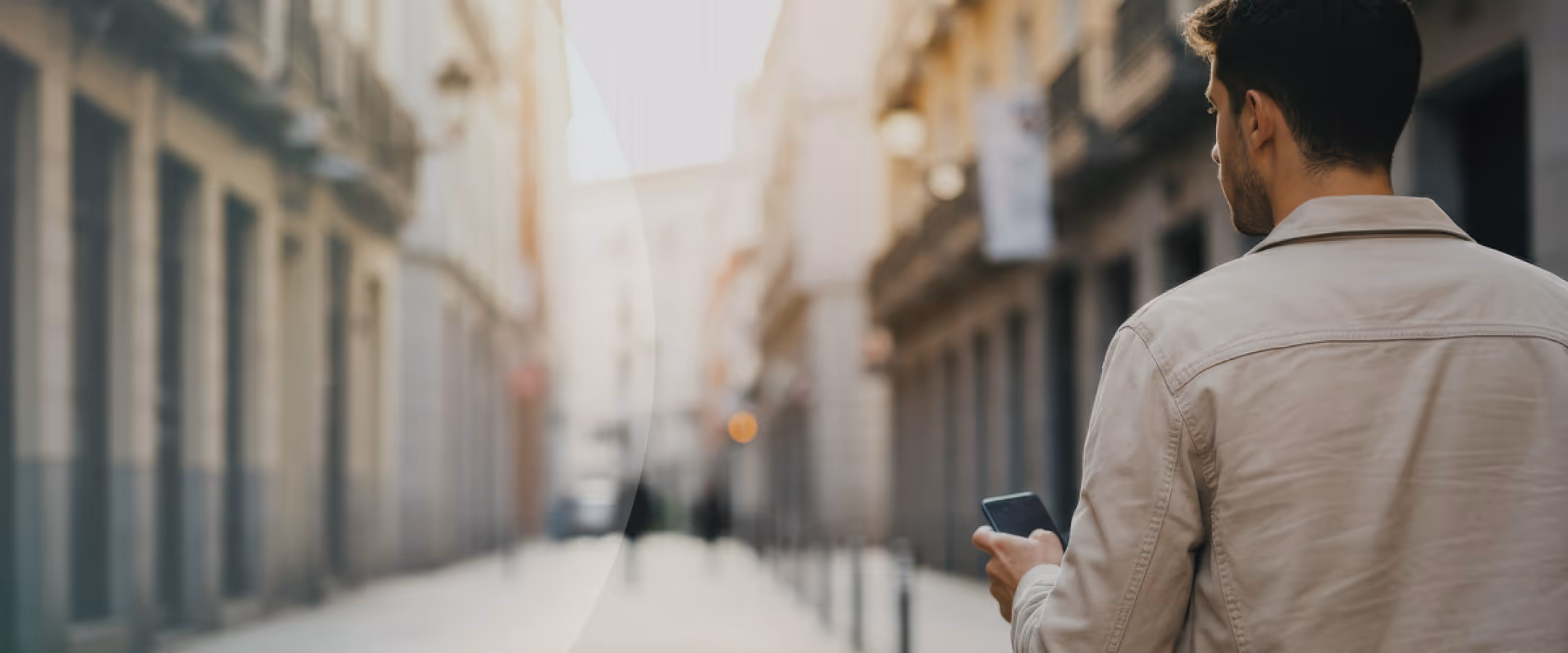 Man in beige jacket looking at his smartphone outdoors with blurred buildings in the background.