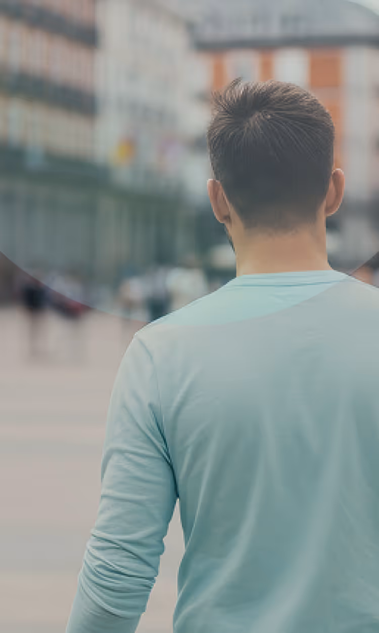 Man wearing a light blue long-sleeve shirt seen from behind, standing on a blurry urban street.