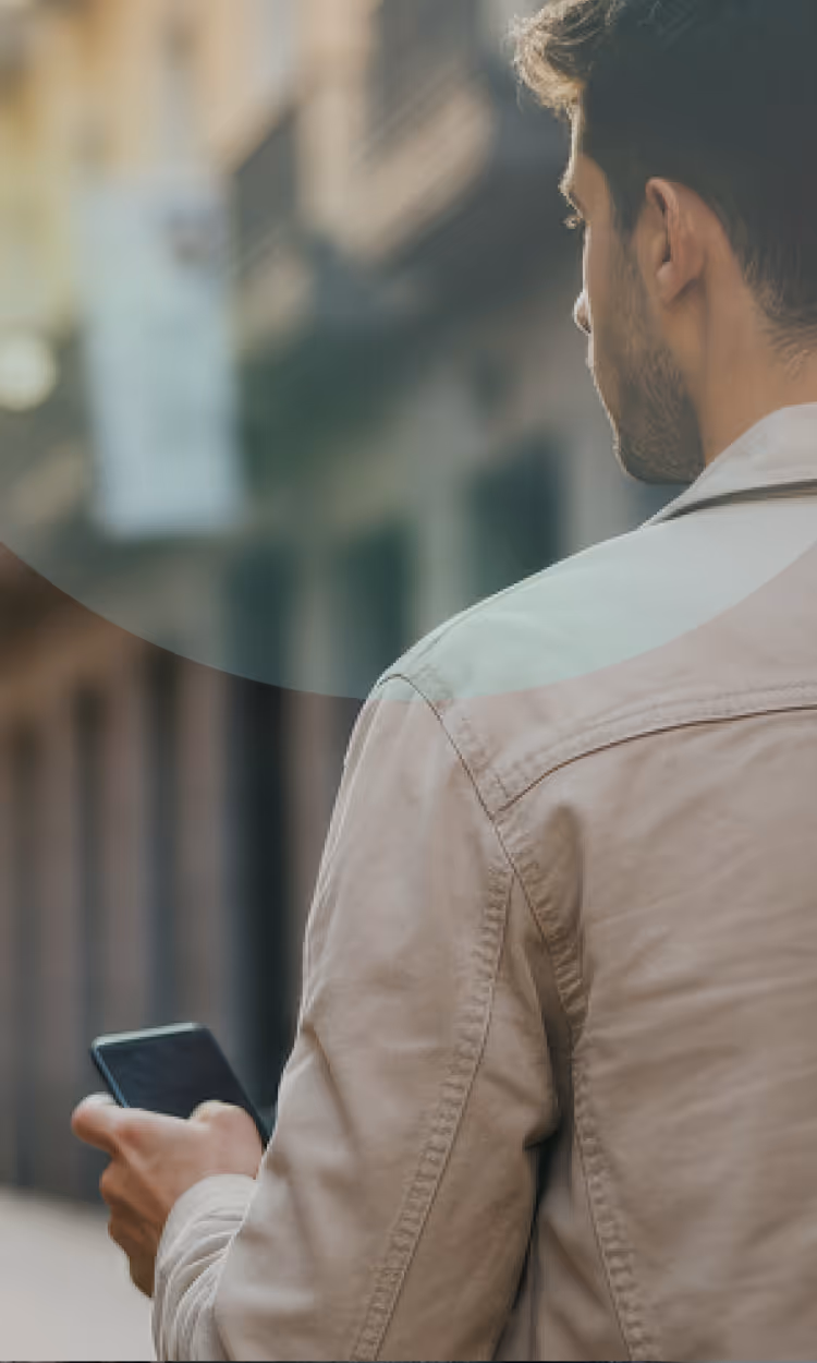 Man in beige jacket looking at his smartphone outdoors with blurred buildings in the background.