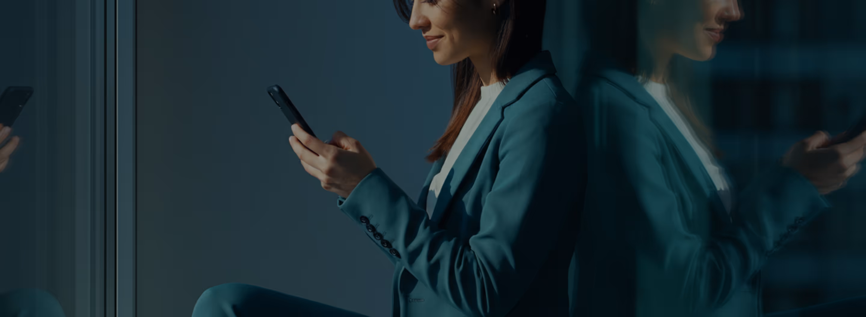 Smiling woman in teal suit sitting by a window looking at her smartphone with its reflection in the glass.