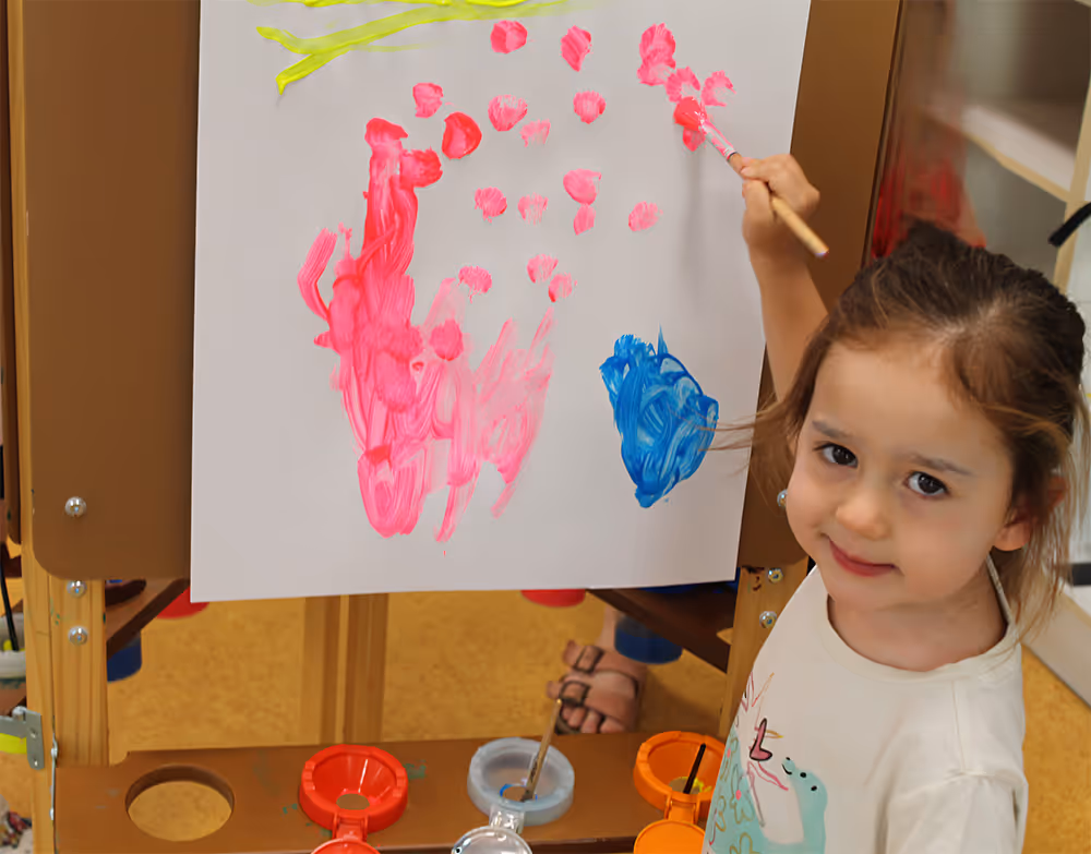 Young girl painting pink and blue colors on a white paper attached to an easel.