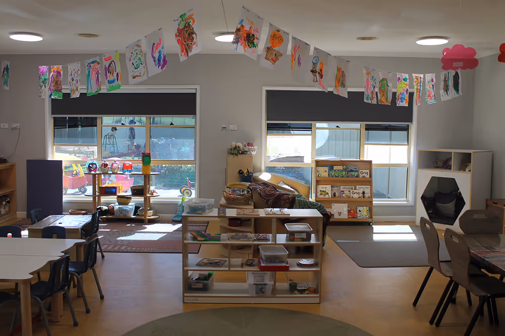 Bright kindergarten classroom with child artwork hanging from the ceiling, small tables and chairs, shelves with toys and books, and large windows letting in natural light.