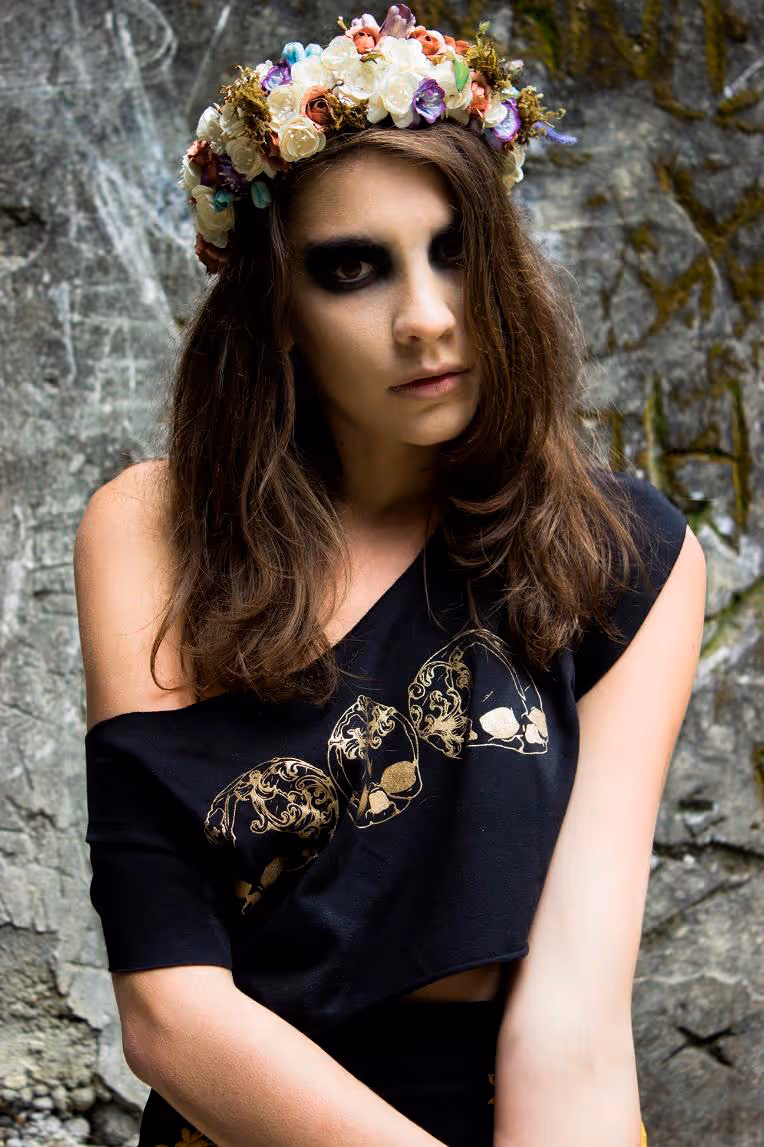 Modelo con corona de flores, con fondo de piedra, mirando hacía la cámara con blusa azul y un hombro al descubierto