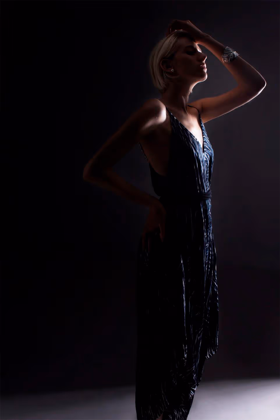 Fotografía de moda con iluminación de silueta que muestra a una mujer rubia posando de perfil con un vestido largo estampado y fondo oscuro.