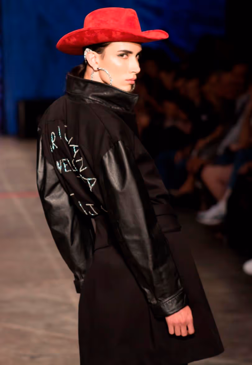 Modelo en pasarela de moda, luciendo sombrero rojo, aretes grande y gabardina negra, volteando a la cámara.