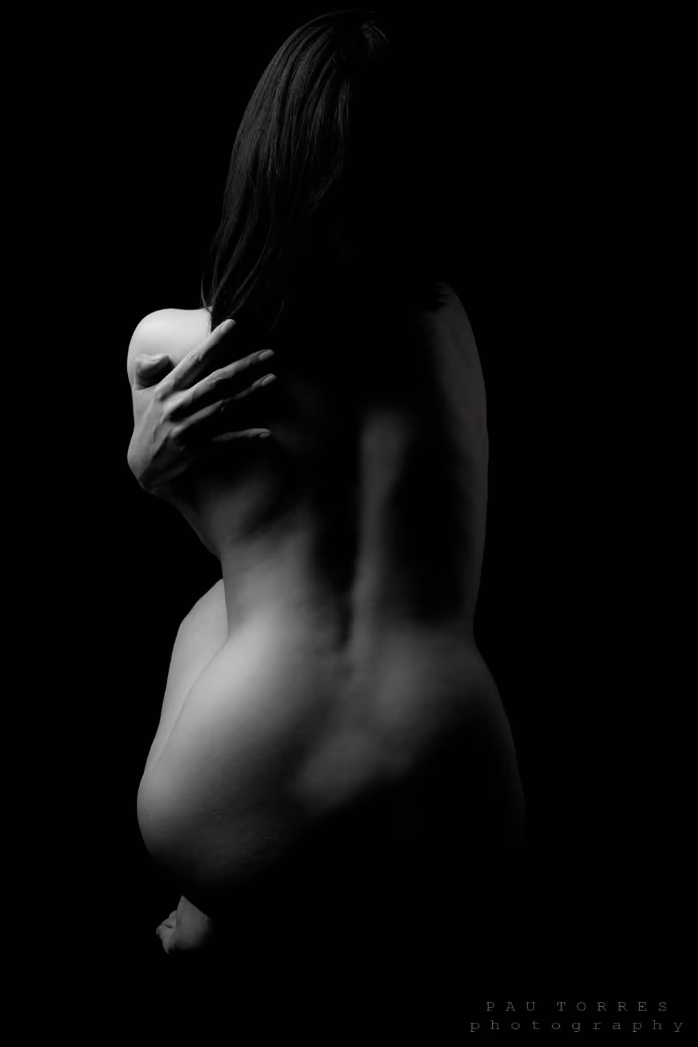 Modelo dando espalda completamente desnuda sentada sobre si misma con fondo oscuro.