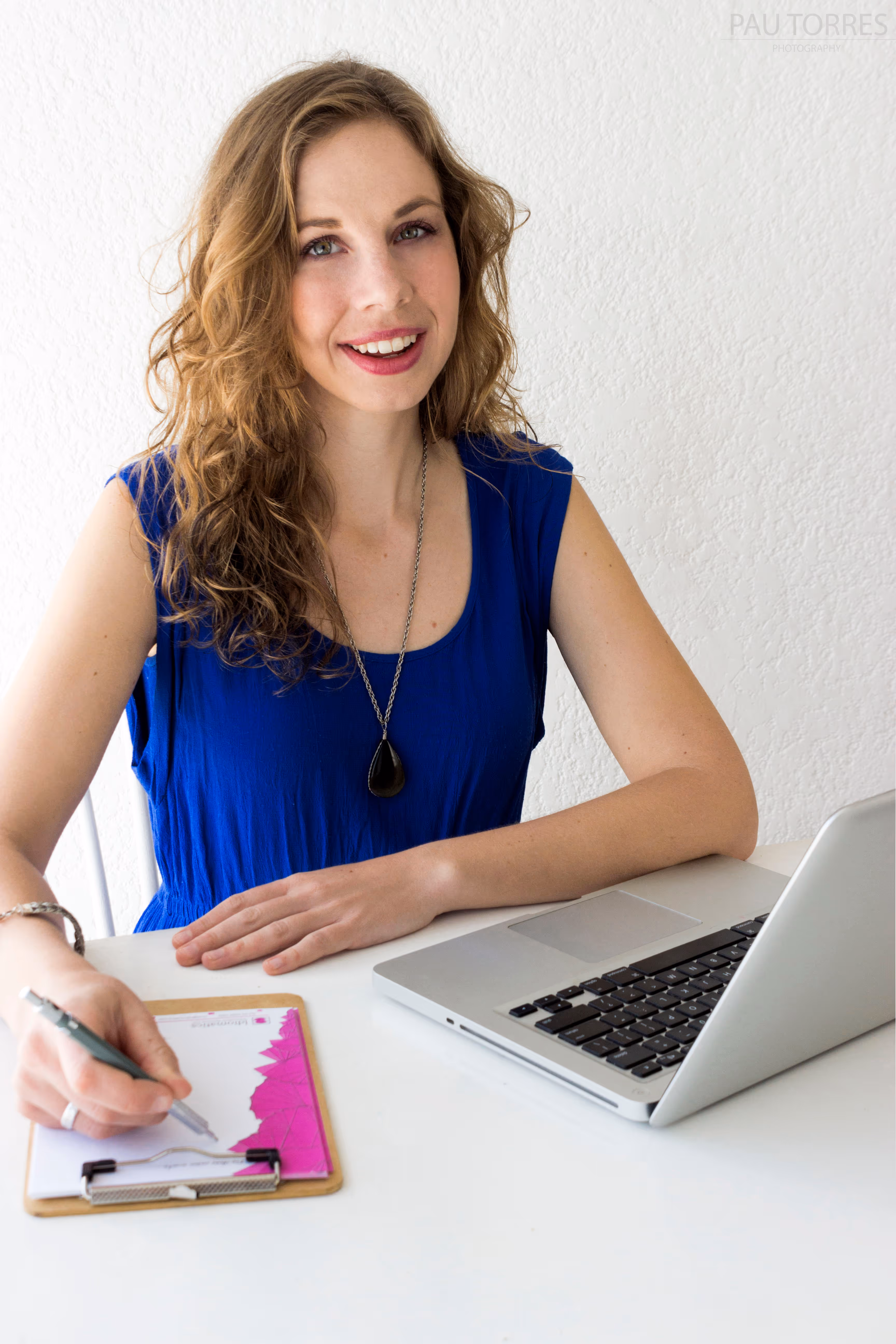 Fotografía de retrato de mujer trabajando en escritorio con laptop, sonriendo a la cámara con fondo de pared blanca.