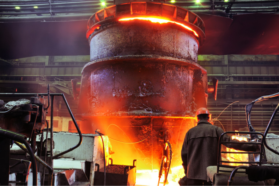 Worker in protective helmet standing near large industrial furnace emitting bright orange molten material inside a factory.