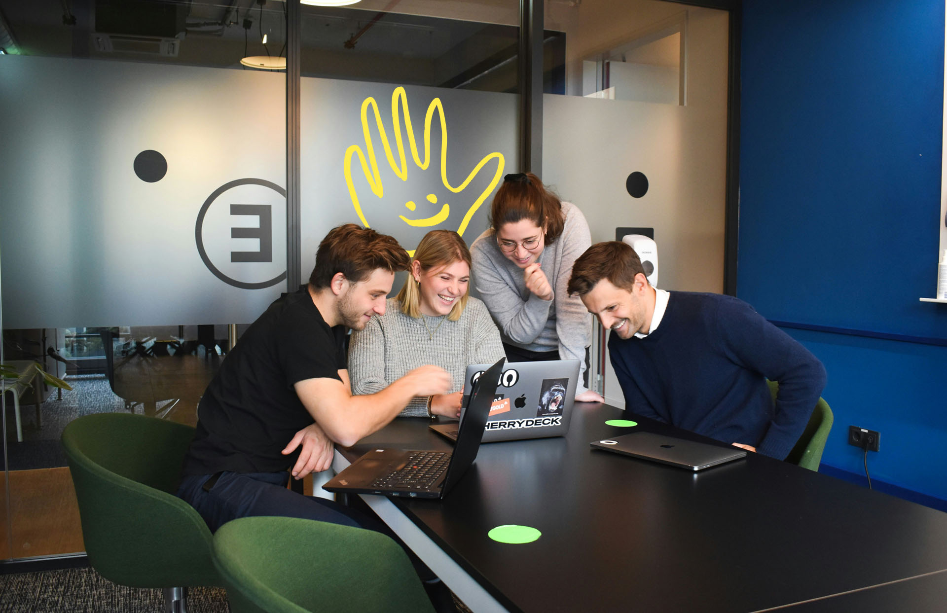 Four colleagues gathered around laptops, smiling and collaborating in a modern office meeting room.