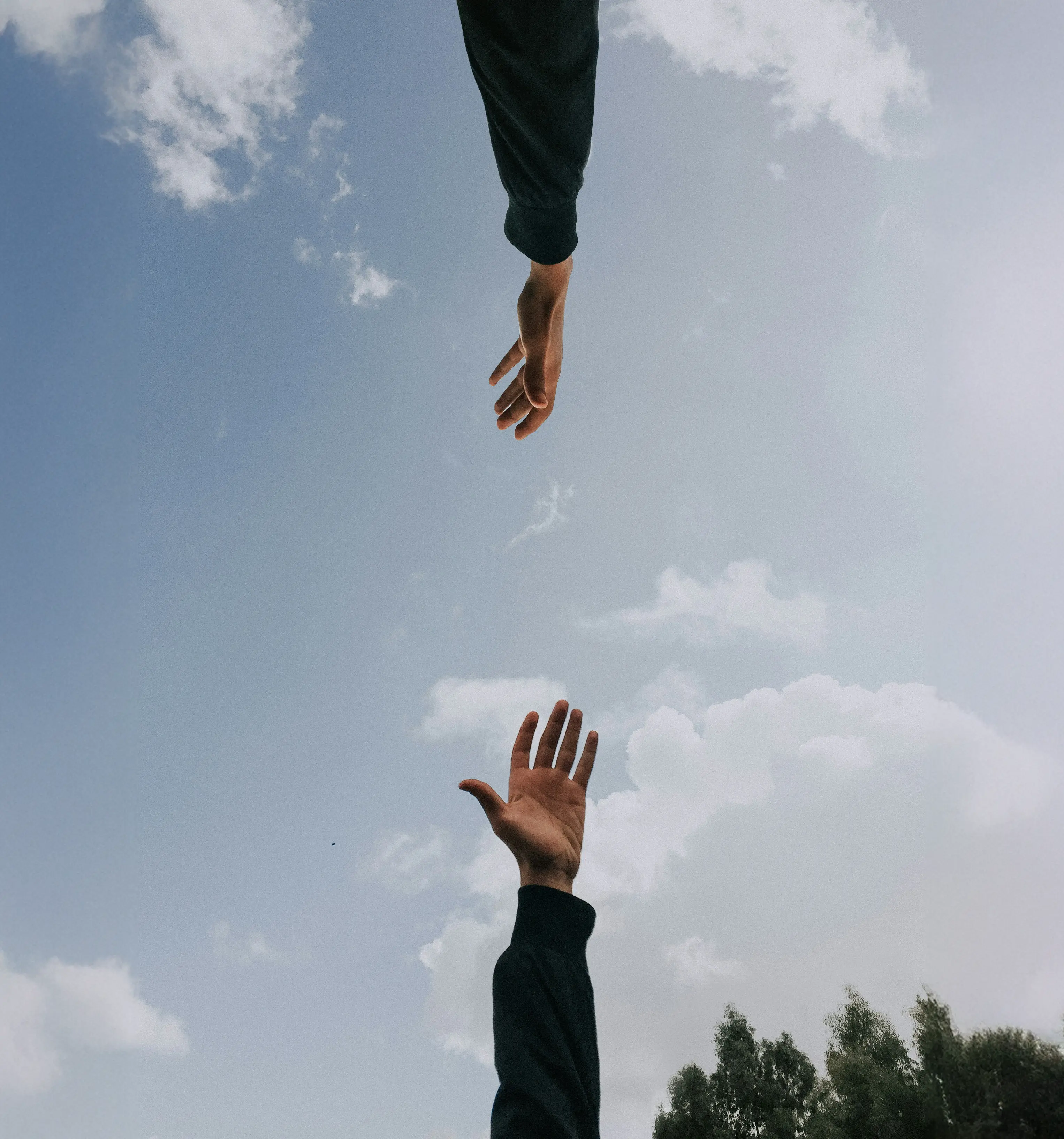 Two hands reaching towards each other against a partly cloudy blue sky with trees below.