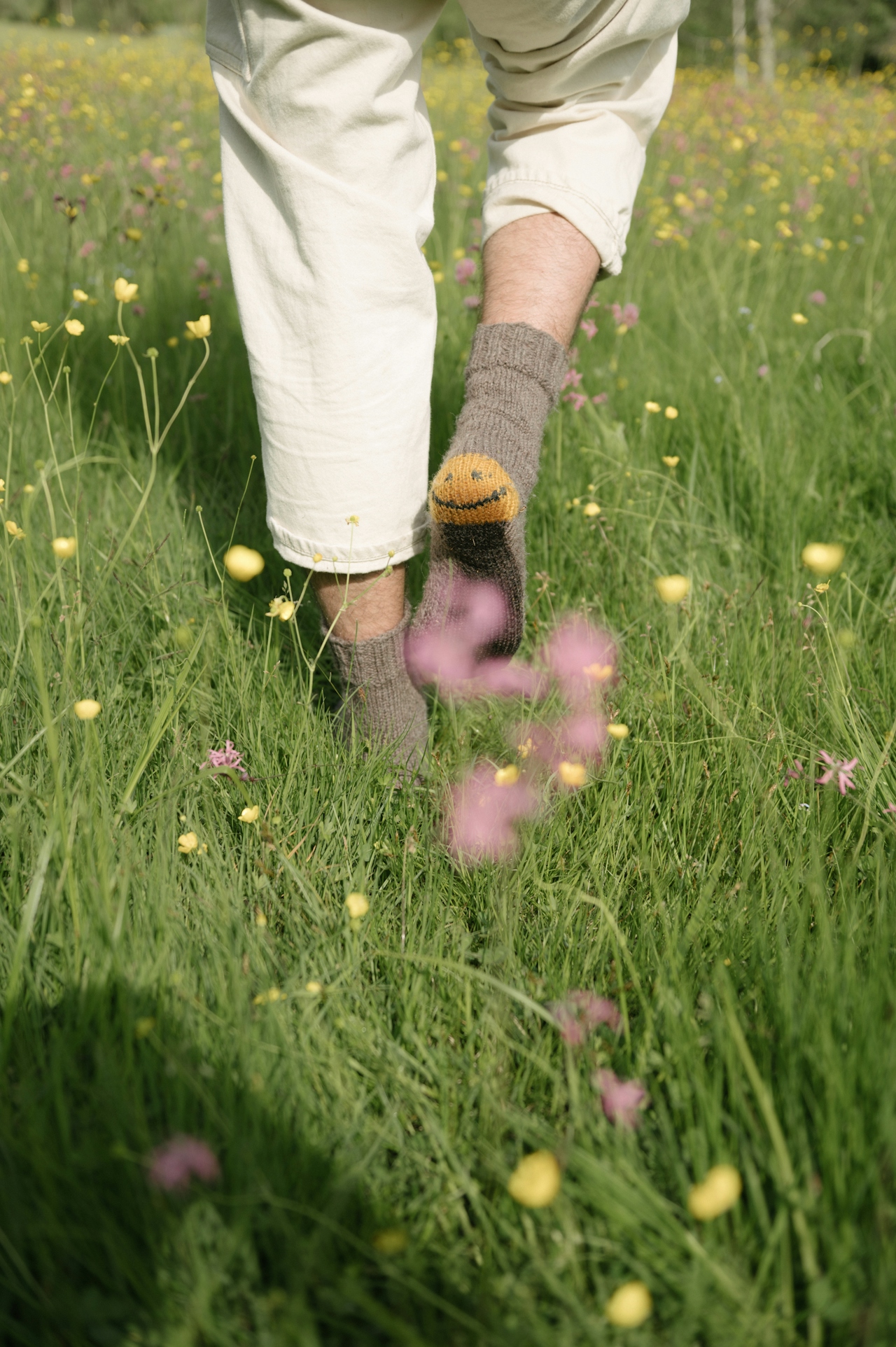 Person walking barefoot in a green field with yellow and pink wildflowers, wearing beige pants and smiley socks.