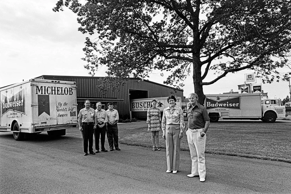 Black and white photo of five people standing in front of a warehouse with Budweiser, Michelob, and Busch beer trucks nearby.