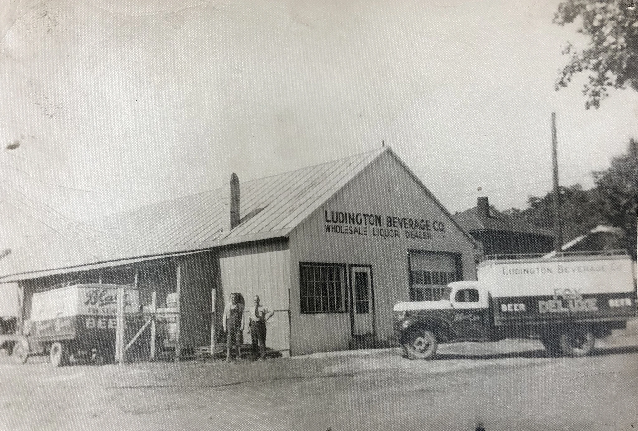 Black-and-white photo of Ludington Beverage Co. warehouse with two delivery trucks and two men standing outside.