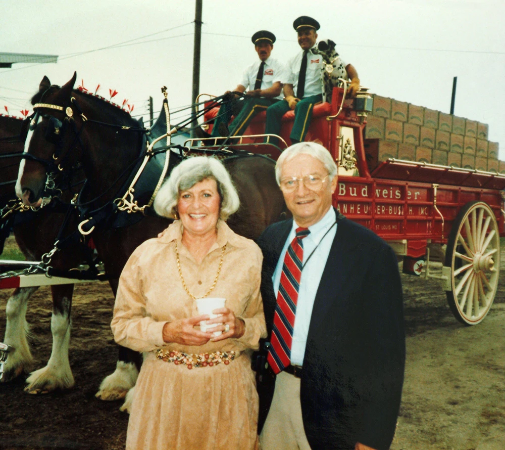 Smiling man and woman standing in front of a red Budweiser horse-drawn wagon with two drivers and a Dalmatian seated on the wagon.