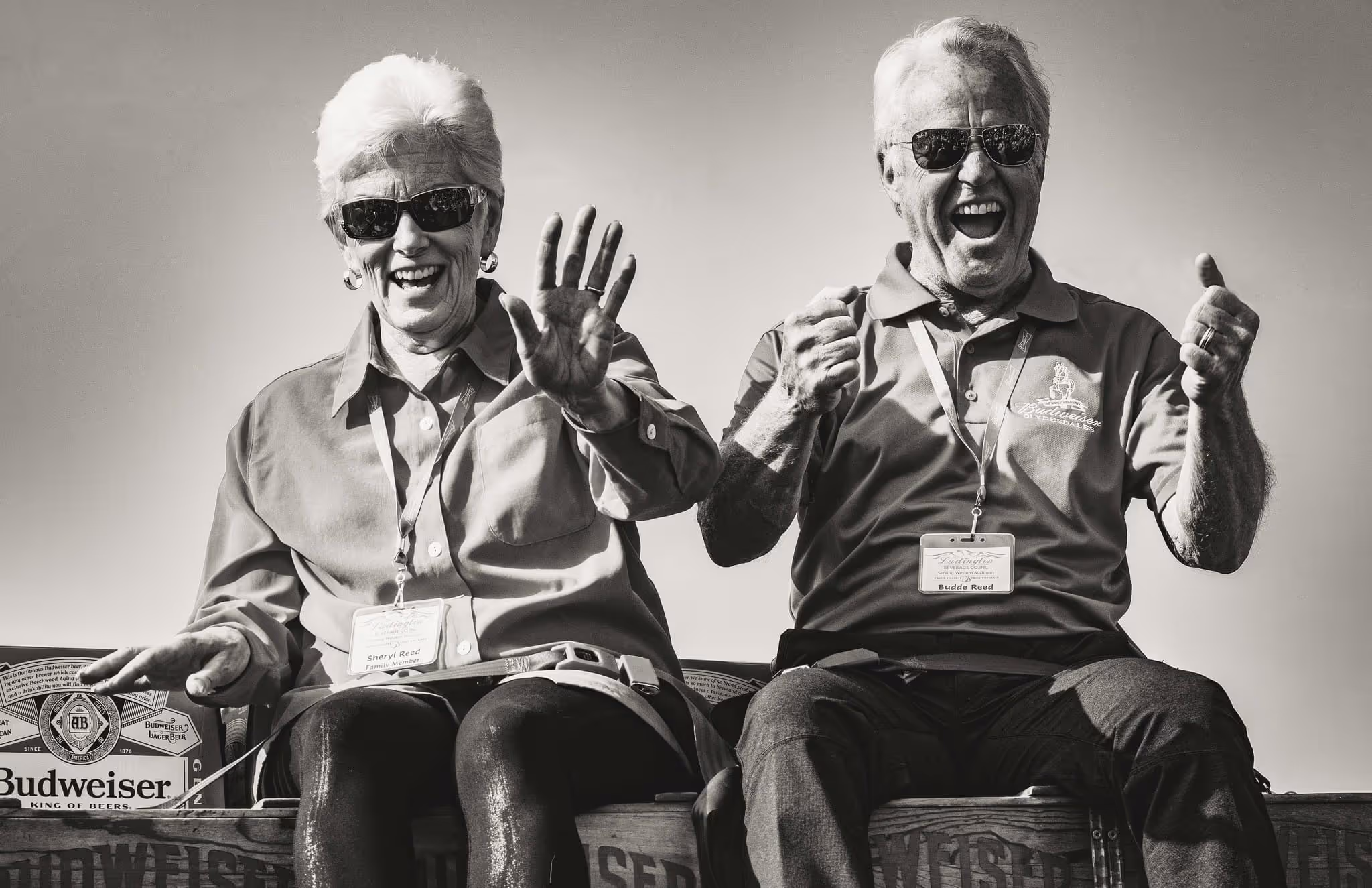 Smiling elderly couple wearing sunglasses and lanyards, sitting on crates with Budweiser branding, celebrating energetically.